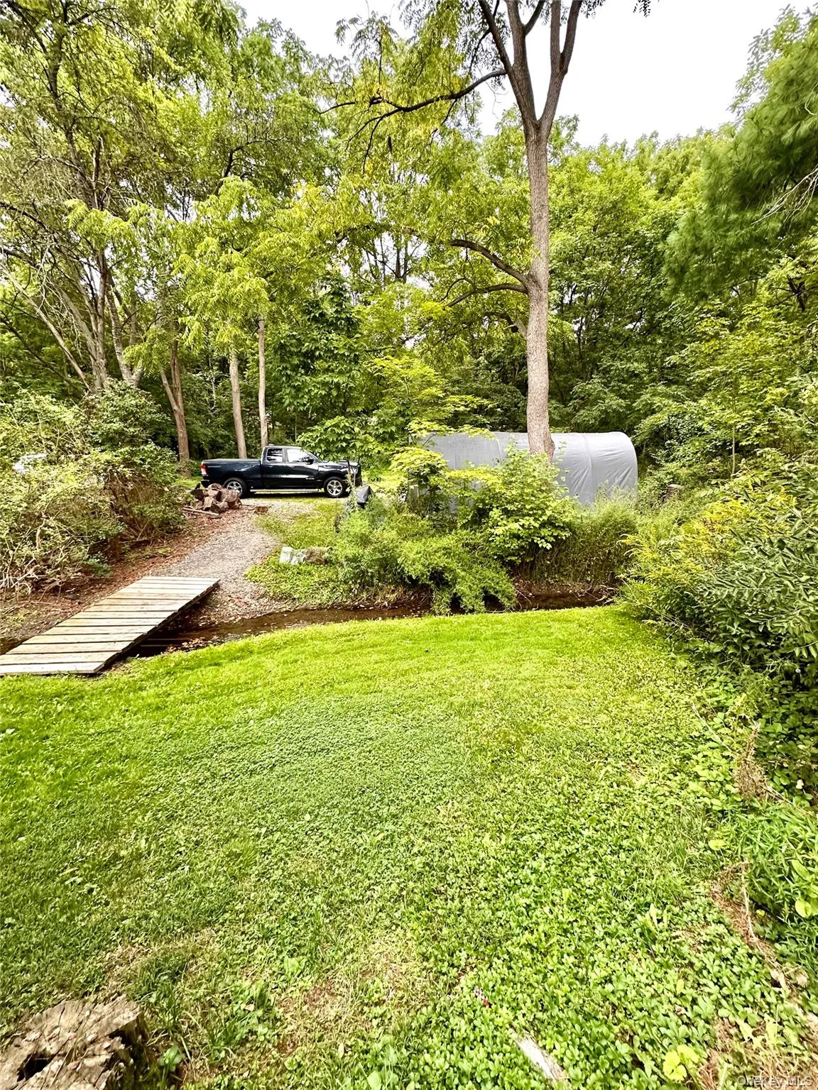 Expansive grassy area with a wooden footbridge over a small water feature, dense mature trees, and a fabric-covered structure Expansive grassy area with a wooden footbridge over a small water feature, dense mature trees, and a fabric-covered structure