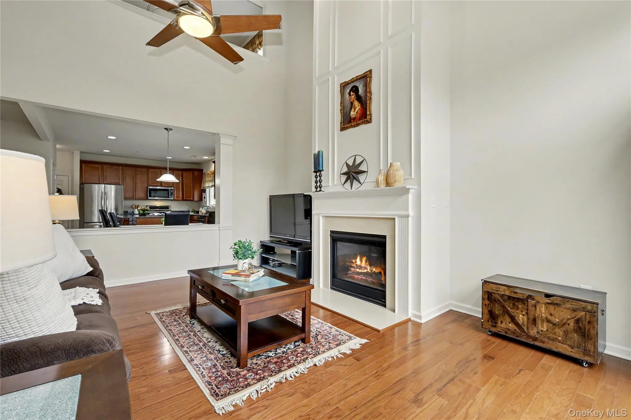 Hardwood flooring, two-story ceiling with a ceiling fan, fireplace with a mantel, and a pass-through opening to the kitchen Hardwood flooring, two-story ceiling with a ceiling fan, fireplace with a mantel, and a pass-through opening to the kitchen