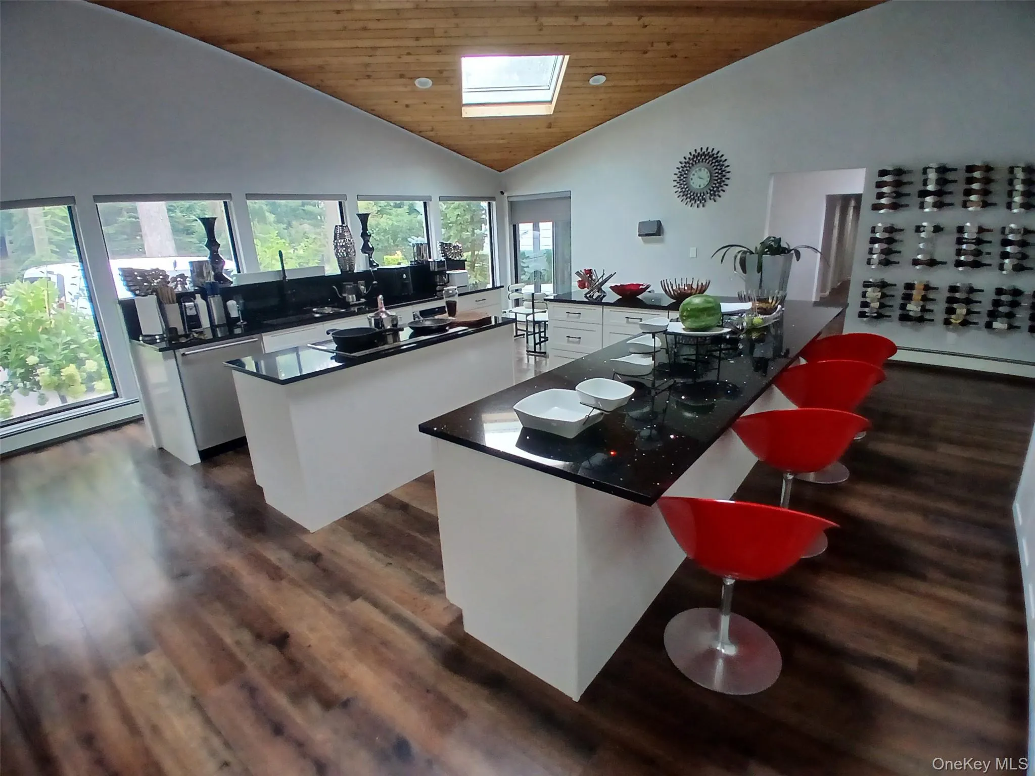 Vaulted wood-finish ceiling with skylight, dark wood-finish flooring, white cabinetry, black countertops, and a built-in wall-mounted wine rack Vaulted wood-finish ceiling with skylight, dark wood-finish flooring, white cabinetry, black countertops, and a built-in wall-mounted wine rack