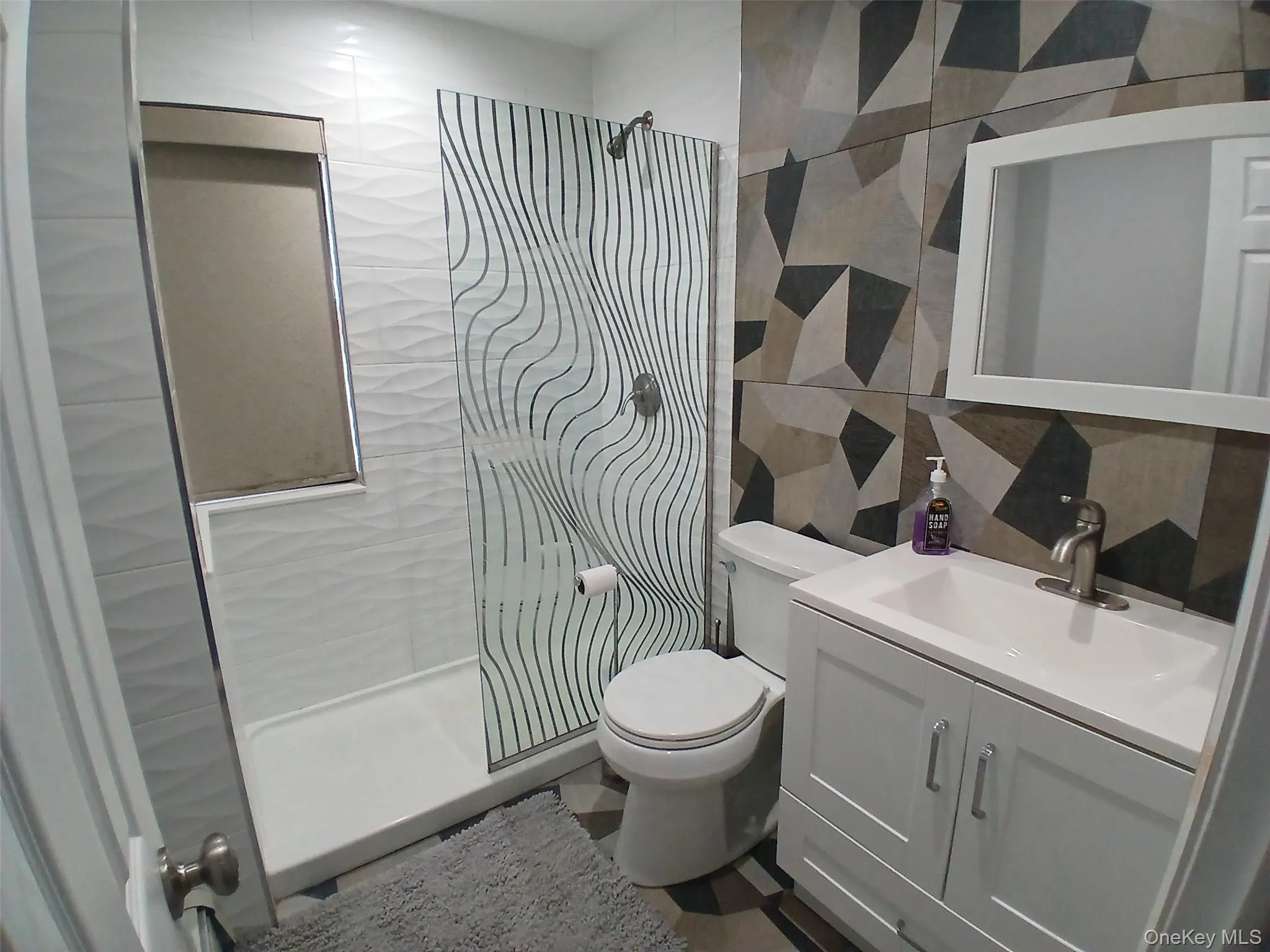 Walk-in shower with patterned glass enclosure, contemporary geometric wall tiles, white vanity with integrated sink, and wall-mounted mirror Walk-in shower with patterned glass enclosure, contemporary geometric wall tiles, white vanity with integrated sink, and wall-mounted mirror