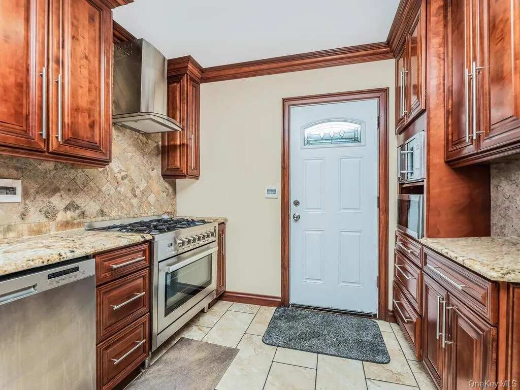 Kitchen with light tile patterned flooring, wall chimney range hood, light stone counters, appliances with stainless steel finishes, and backsplash Kitchen with light tile patterned flooring, wall chimney range hood, light stone counters, appliances with stainless steel finishes, and backsplash