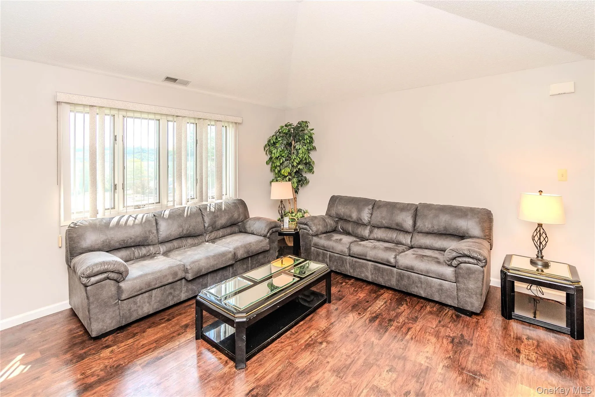 Vaulted ceiling, wood-finish flooring, large window with vertical blinds, neutral wall paint Vaulted ceiling, wood-finish flooring, large window with vertical blinds, neutral wall paint