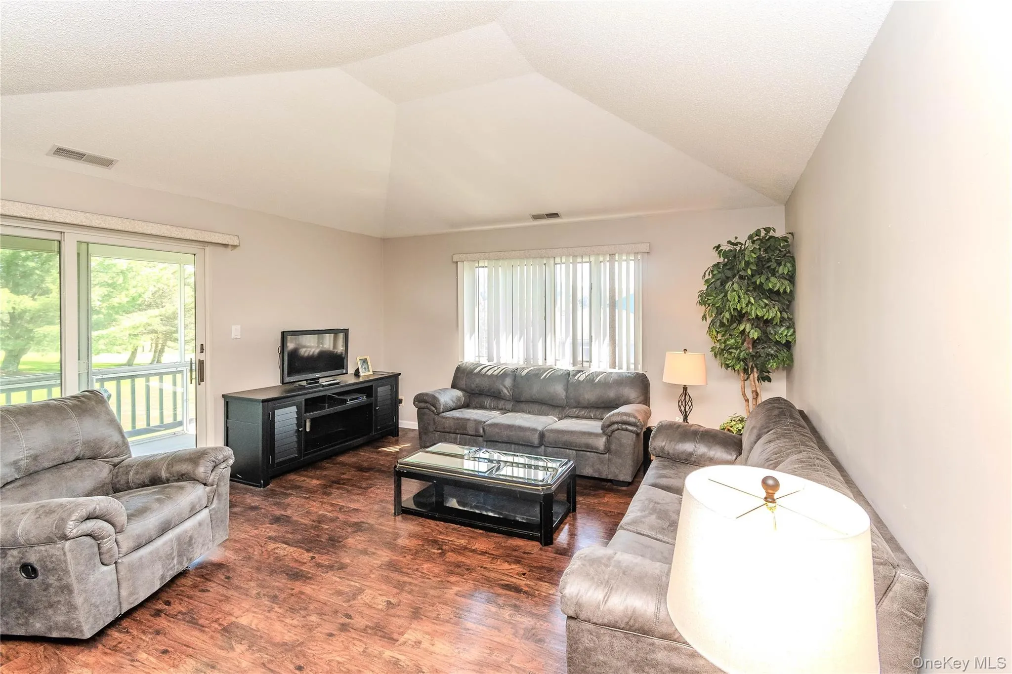 Vaulted ceiling living area with wood-finish flooring, sliding glass door with balcony access, and natural light from window treatments Vaulted ceiling living area with wood-finish flooring, sliding glass door with balcony access, and natural light from window treatments