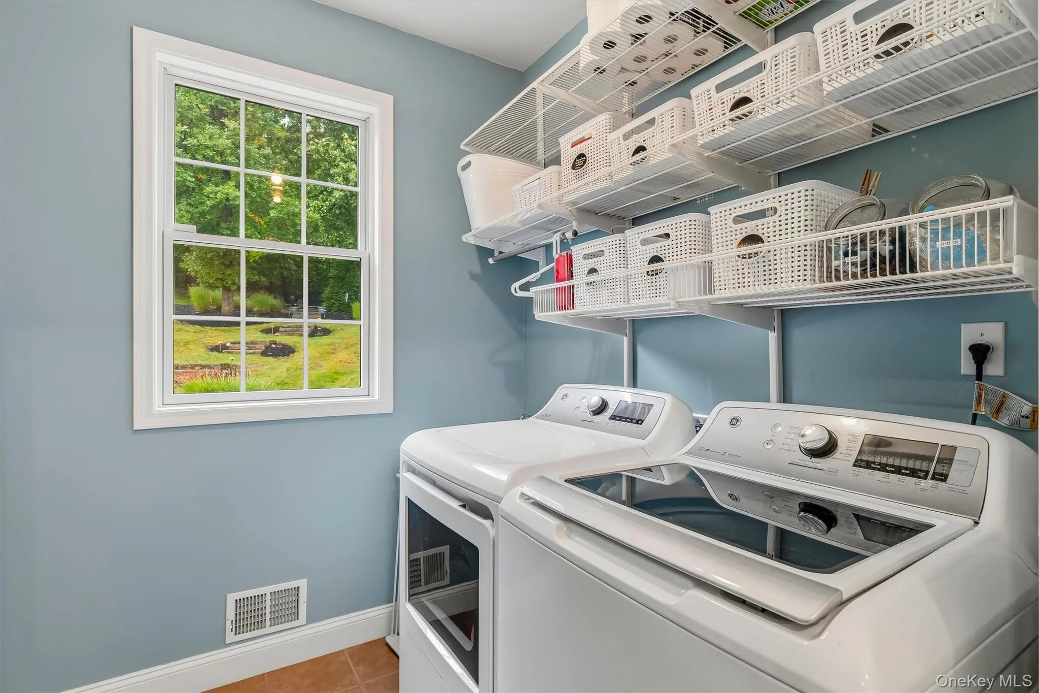 This bright and efficiently designed laundry room makes everyday chores feel effortless. A large window fills the space with natural light and offers a rear view of the beautifully landscaped yard, while extensive wall-mounted shelving provides exceptional storage for laundry supplies, baskets, and household essentials. With side-by-side washer and dryer placement, durable tile flooring, and a fresh, cheerful color palette, this well-organized room blends practicality, convenience, and style. This bright and efficiently designed laundry room makes everyday chores feel effortless. A large window fills the space with natural light and offers a rear view of the beautifully landscaped yard, while extensive wall-mounted shelving provides exceptional storage for laundry supplies, baskets, and household essentials. With side-by-side washer and dryer placement, durable tile flooring, and a fresh, cheerful color palette, this well-organized room blends practicality, convenience, and style.
