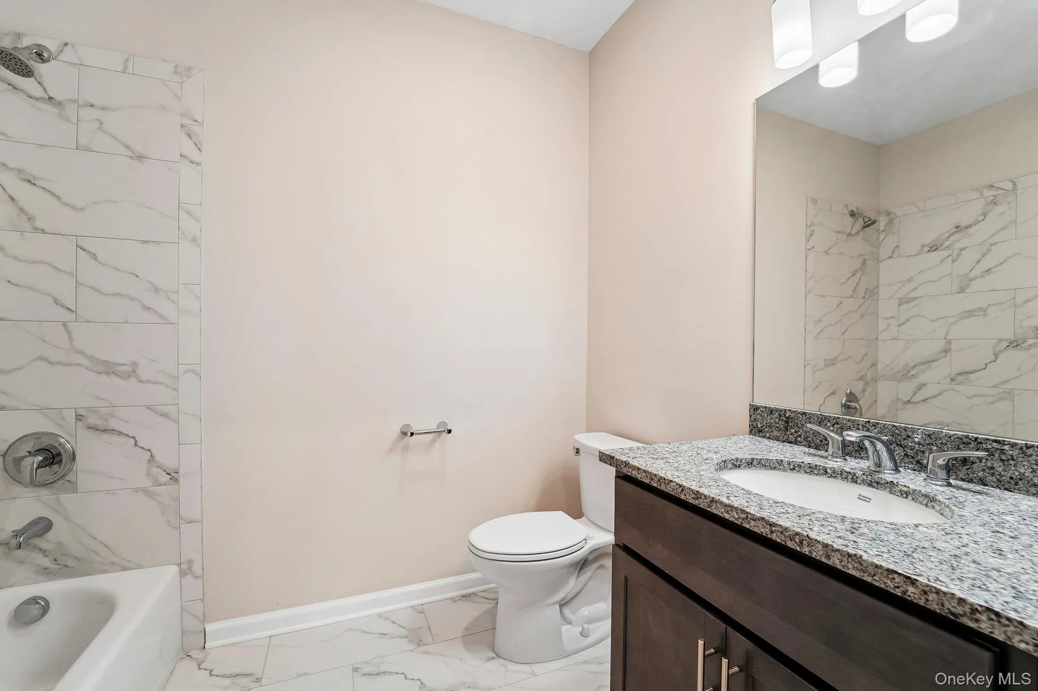 Bathroom featuring white marble-look floor and shower wall tile, a dark wood-finish vanity with a granite countertop, and chrome fixtures Bathroom featuring white marble-look floor and shower wall tile, a dark wood-finish vanity with a granite countertop, and chrome fixtures