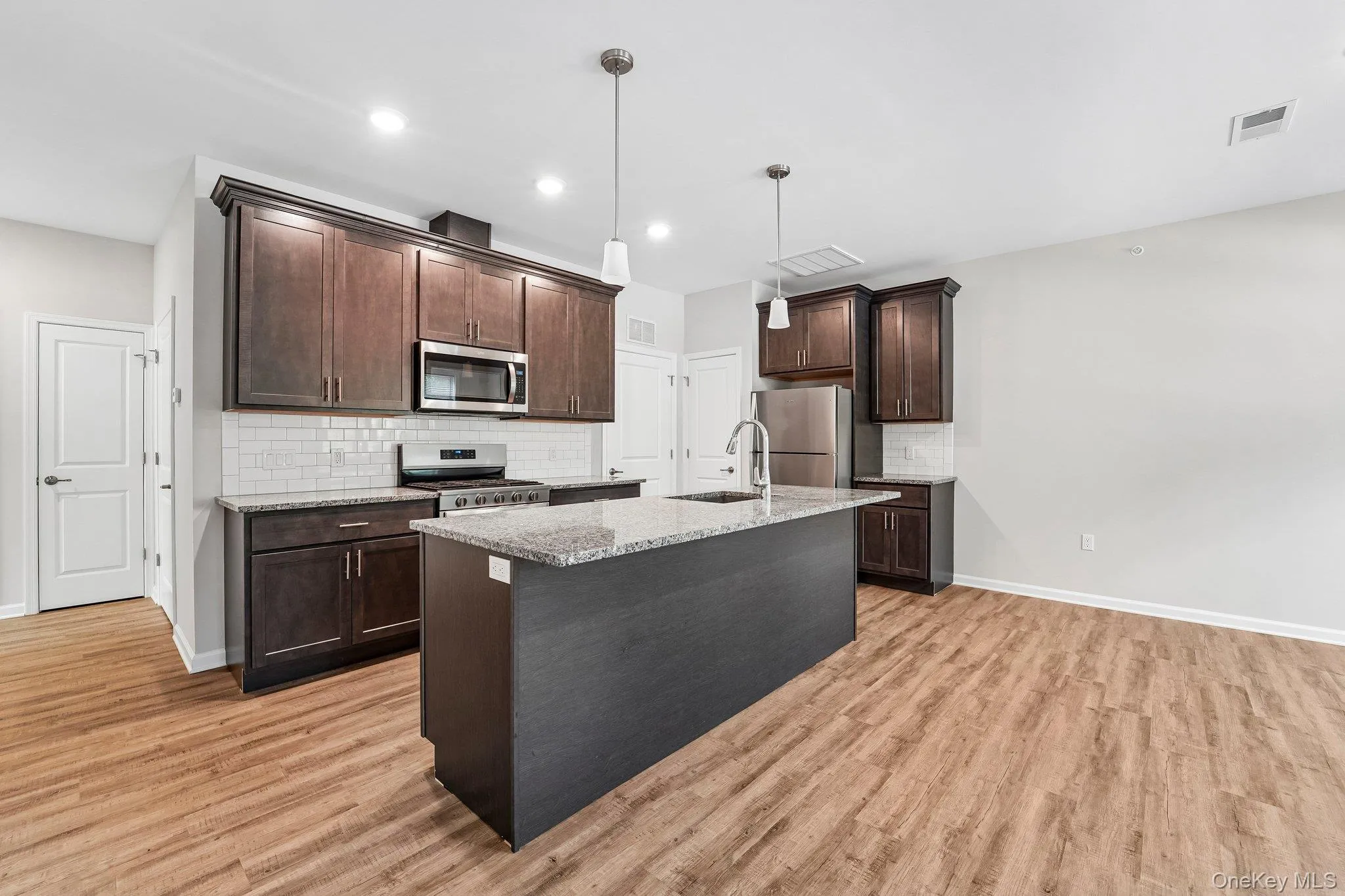 Wood-finish flooring throughout, dark wood cabinetry, stainless steel appliances, white subway tile backsplash, and a kitchen island with pendant lighting Wood-finish flooring throughout, dark wood cabinetry, stainless steel appliances, white subway tile backsplash, and a kitchen island with pendant lighting