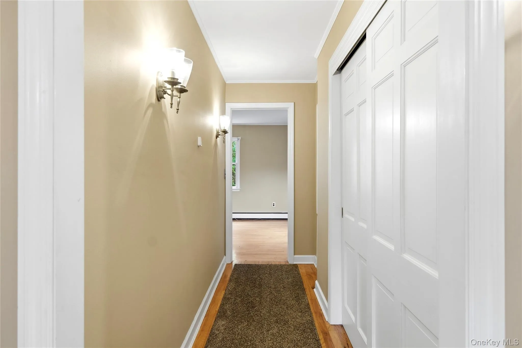 Hallway leading to primary en suite .Hardwood flooring with a runner, sconce lighting, and a white paneled bi-fold closet door Hallway leading to primary en suite .Hardwood flooring with a runner, sconce lighting, and a white paneled bi-fold closet door