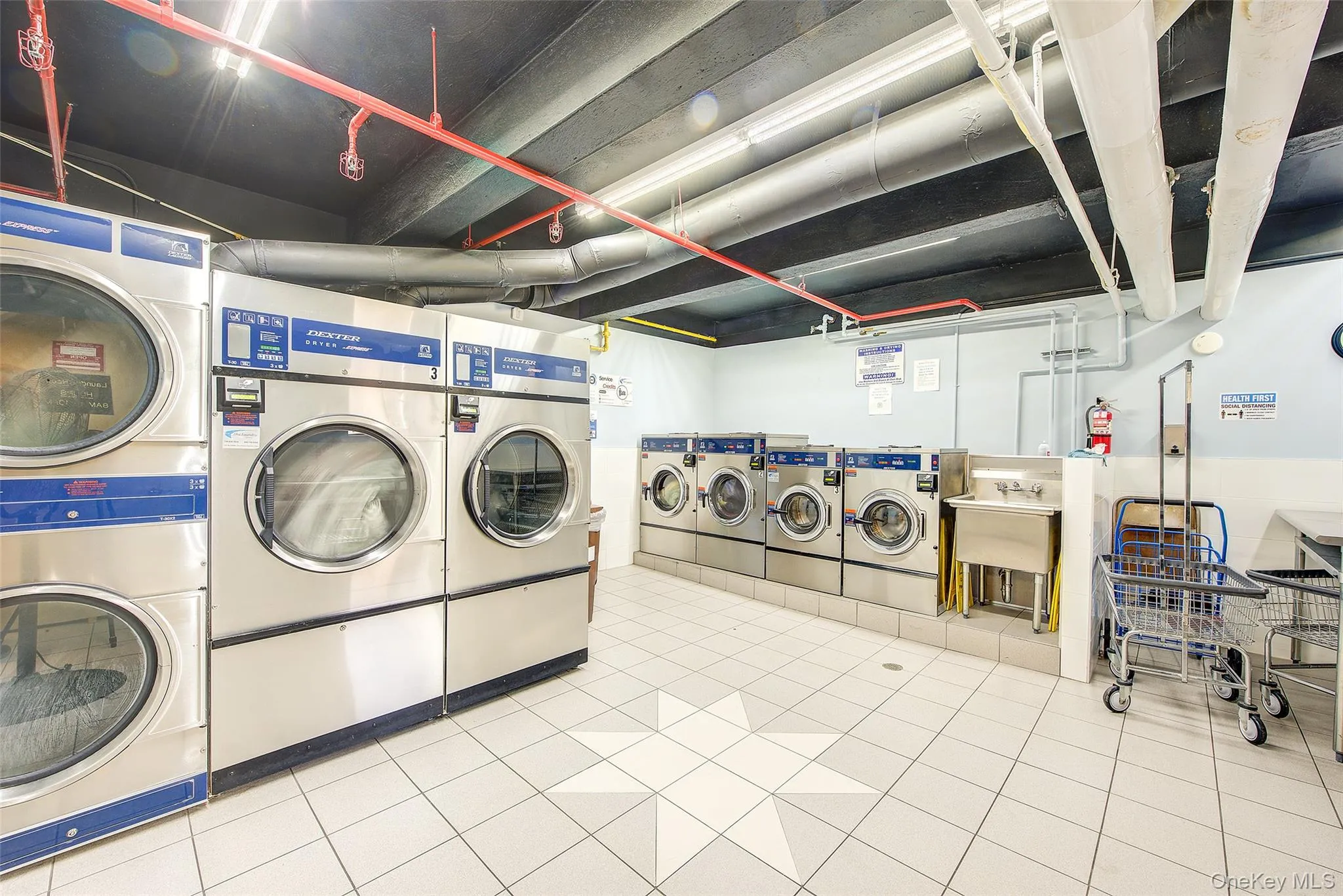 Commercial-grade laundry appliances with tile flooring and exposed ceiling utilities Commercial-grade laundry appliances with tile flooring and exposed ceiling utilities