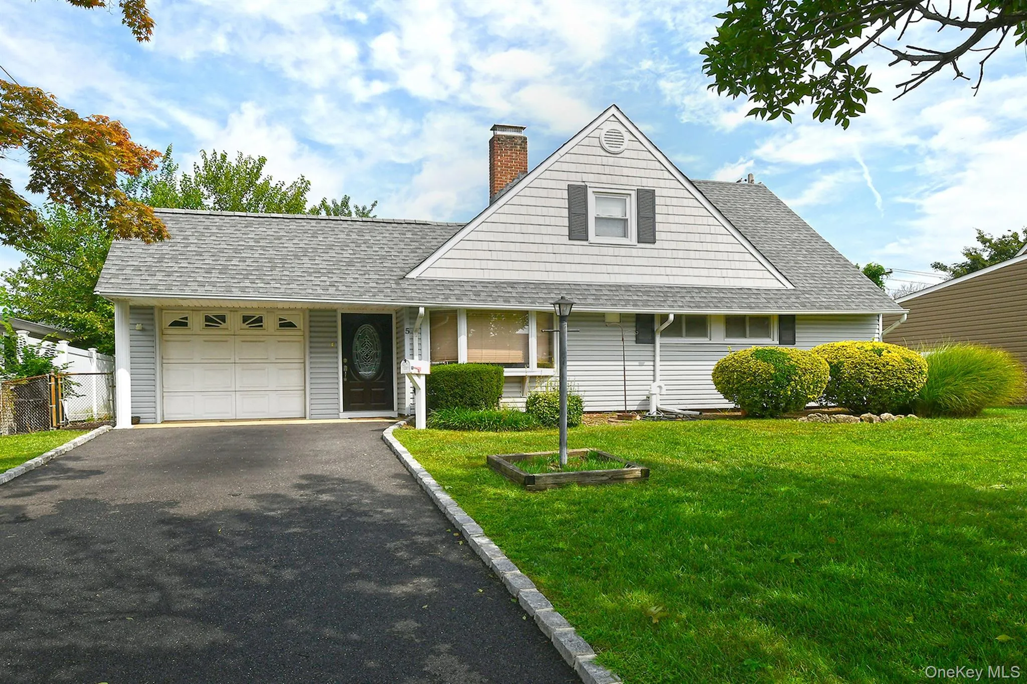 Gable roofline with shingles, attached garage, paved driveway with stone border, and a well-maintained lawn with mature shrubbery Gable roofline with shingles, attached garage, paved driveway with stone border, and a well-maintained lawn with mature shrubbery