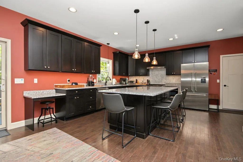 Kitchen featuring dark wood-finish cabinetry, light-toned countertops, a central island with pendant lighting, a stainless steel refrigerator, and wood-finish flooring Kitchen featuring dark wood-finish cabinetry, light-toned countertops, a central island with pendant lighting, a stainless steel refrigerator, and wood-finish flooring