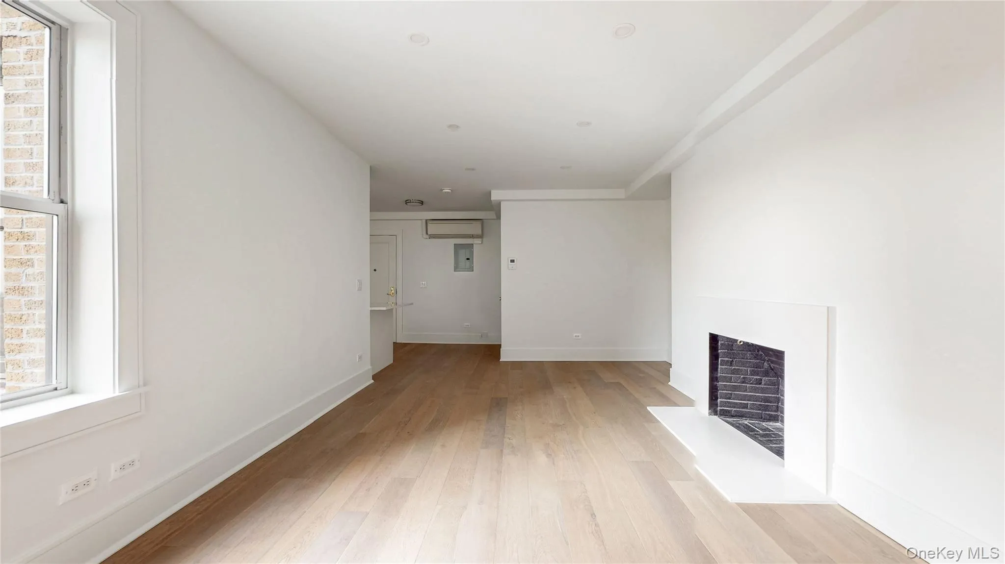 Light wood-finish flooring, recessed lighting, and a modern fireplace with a white mantel and black brick interior Light wood-finish flooring, recessed lighting, and a modern fireplace with a white mantel and black brick interior
