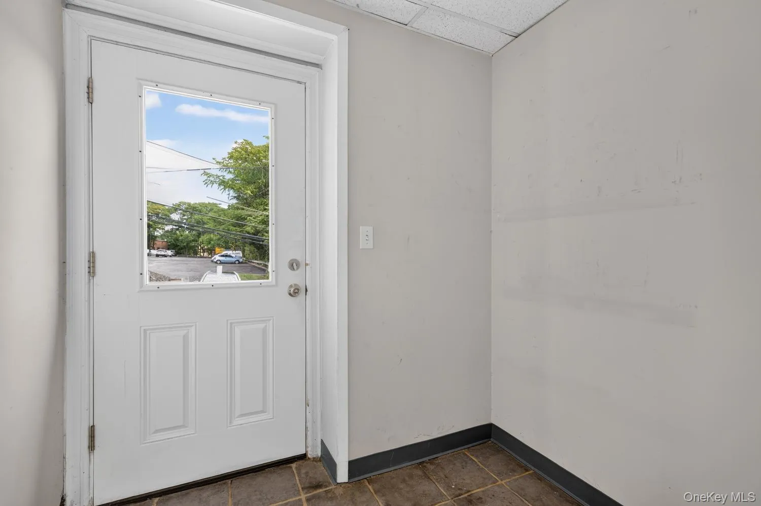 White paneled door with integrated window and chrome hardware, set against light gray walls with dark baseboards, and brown tile flooring White paneled door with integrated window and chrome hardware, set against light gray walls with dark baseboards, and brown tile flooring