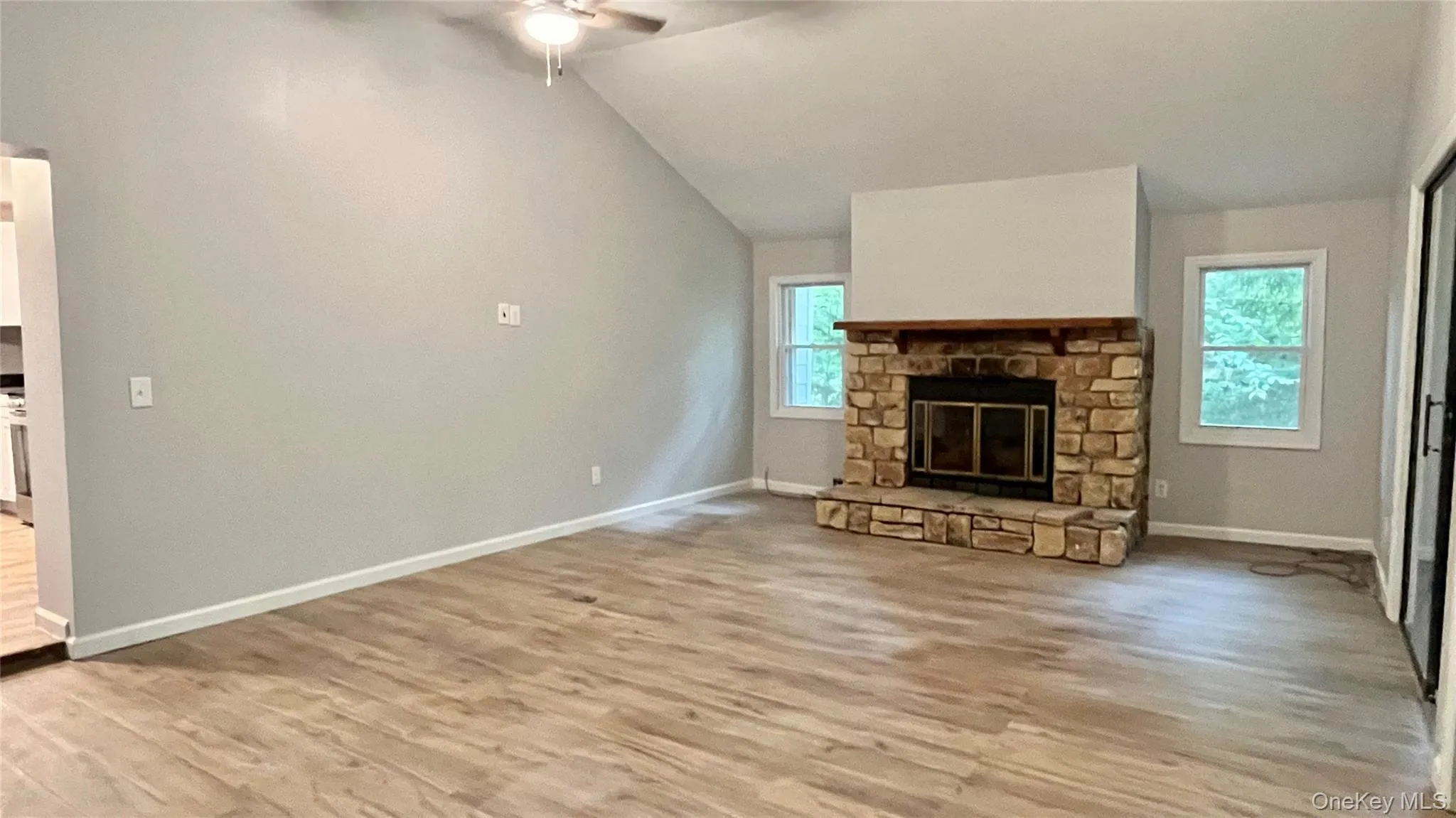 Vaulted ceiling with ceiling fan, stone-clad fireplace with wood mantel, wood-finish flooring, and multiple windows Vaulted ceiling with ceiling fan, stone-clad fireplace with wood mantel, wood-finish flooring, and multiple windows