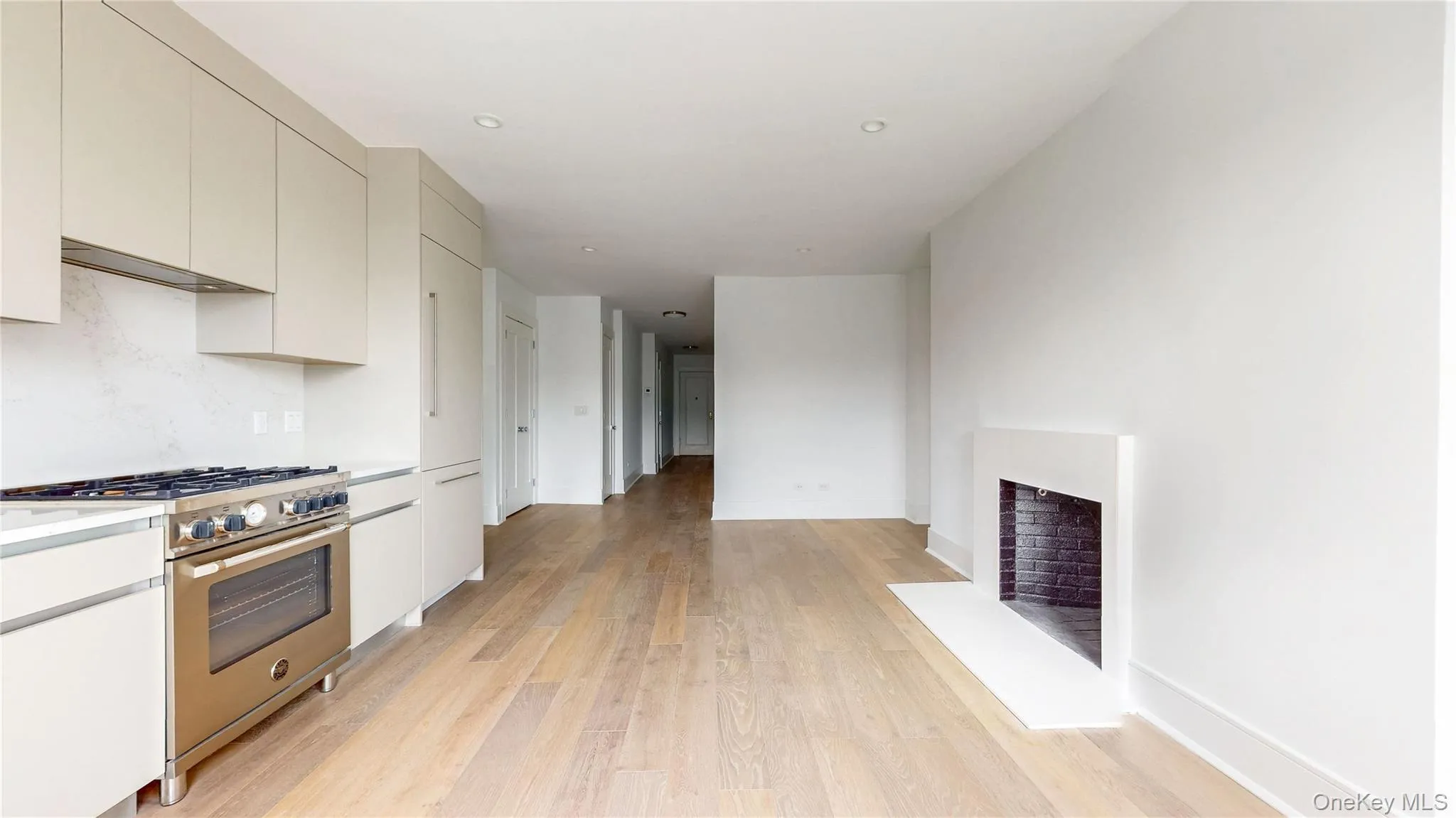 Integrated kitchen cabinetry with stainless steel range, light-toned wood-finish flooring, recessed ceiling lighting, and a modern fireplace with a dark brick interior Integrated kitchen cabinetry with stainless steel range, light-toned wood-finish flooring, recessed ceiling lighting, and a modern fireplace with a dark brick interior