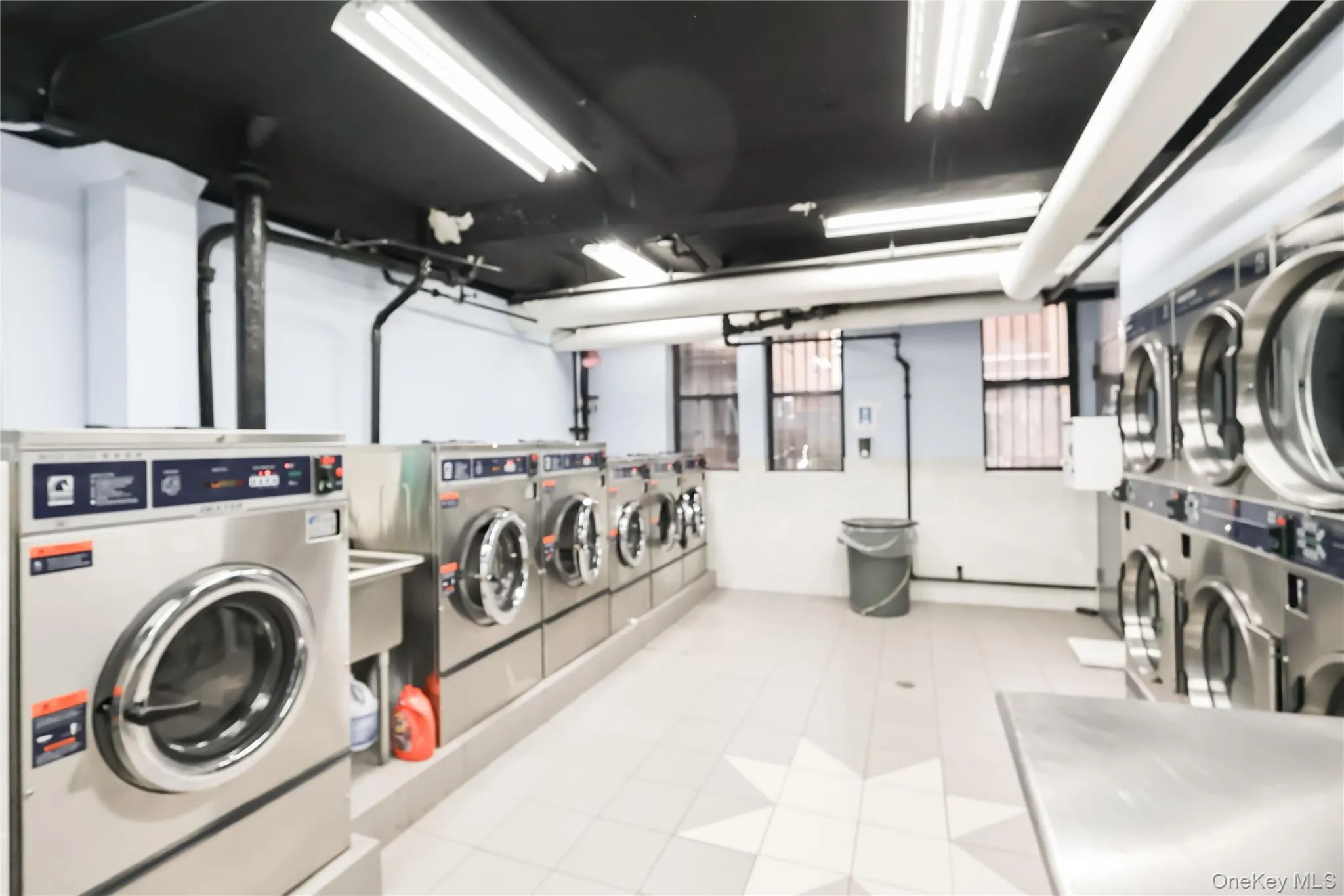 Equipped laundry room with multiple stainless steel washing machines and dryers, tile flooring, and utility sink Equipped laundry room with multiple stainless steel washing machines and dryers, tile flooring, and utility sink
