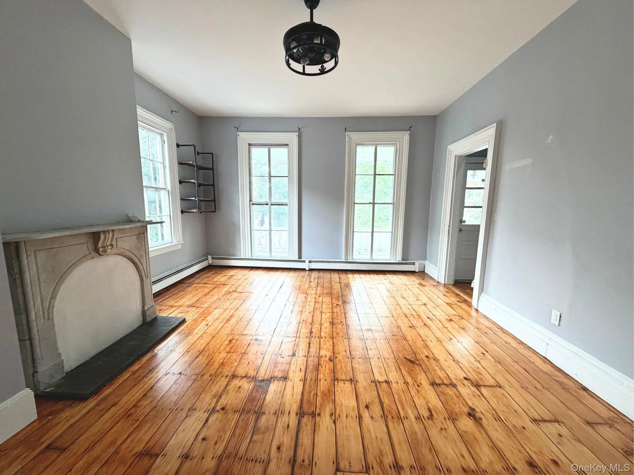 Hardwood flooring throughout, faux fireplace with dark hearth, two large windows, built-in wall shelving, and a ceiling-mounted light fixture Hardwood flooring throughout, faux fireplace with dark hearth, two large windows, built-in wall shelving, and a ceiling-mounted light fixture