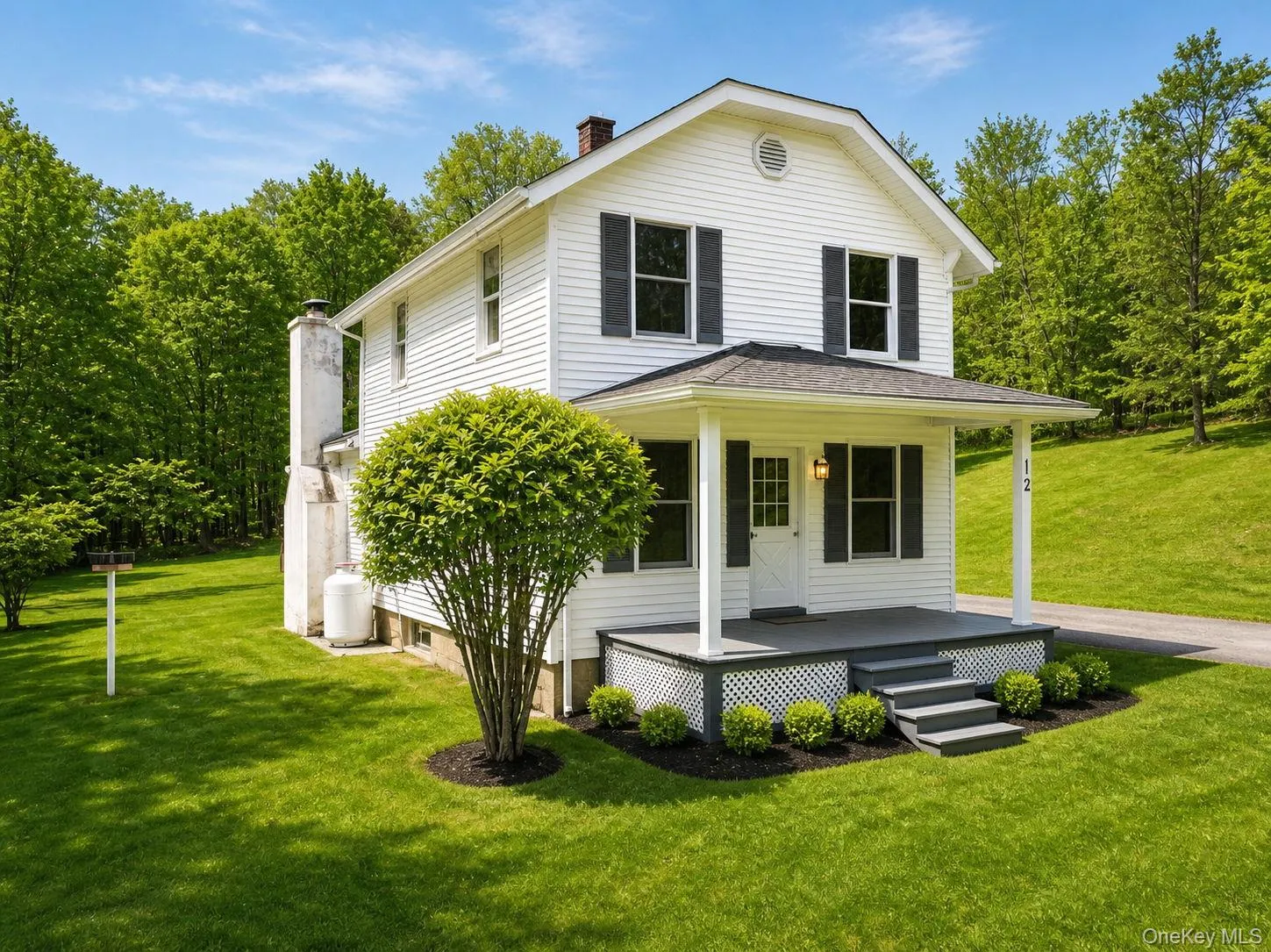Two-story residence featuring white siding, dark shutters, a covered porch with steps, a manicured lawn, and surrounding mature trees Two-story residence featuring white siding, dark shutters, a covered porch with steps, a manicured lawn, and surrounding mature trees