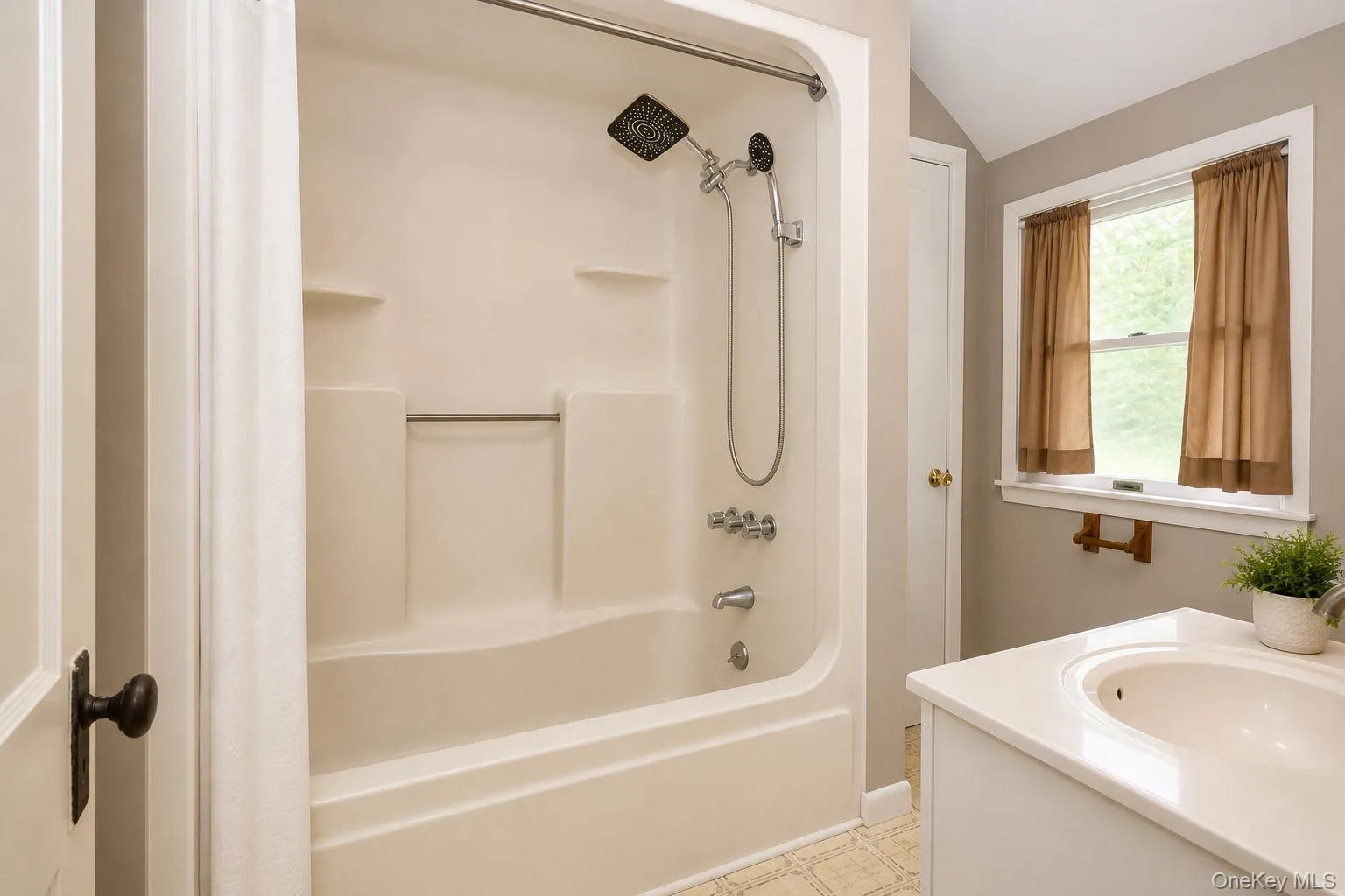 Bathroom featuring a single-piece tub and shower unit with an adjustable showerhead, a single vanity with an integrated sink, and an overhead window Bathroom featuring a single-piece tub and shower unit with an adjustable showerhead, a single vanity with an integrated sink, and an overhead window