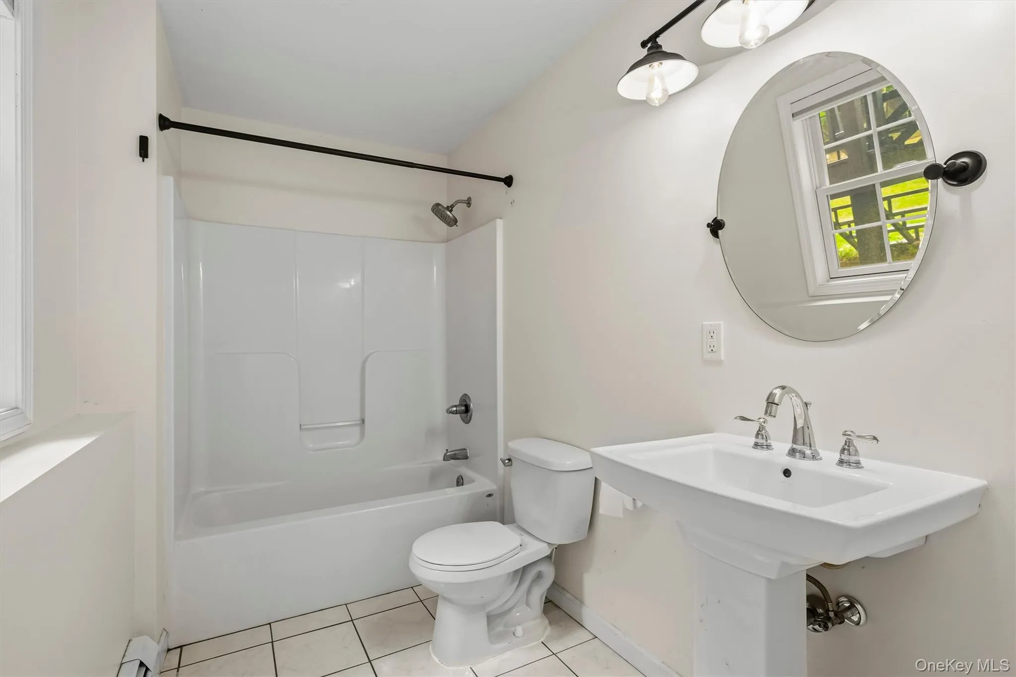White pedestal sink with chrome faucet, oval mirror, track lighting, white tile flooring, and a white tub/shower combination White pedestal sink with chrome faucet, oval mirror, track lighting, white tile flooring, and a white tub/shower combination