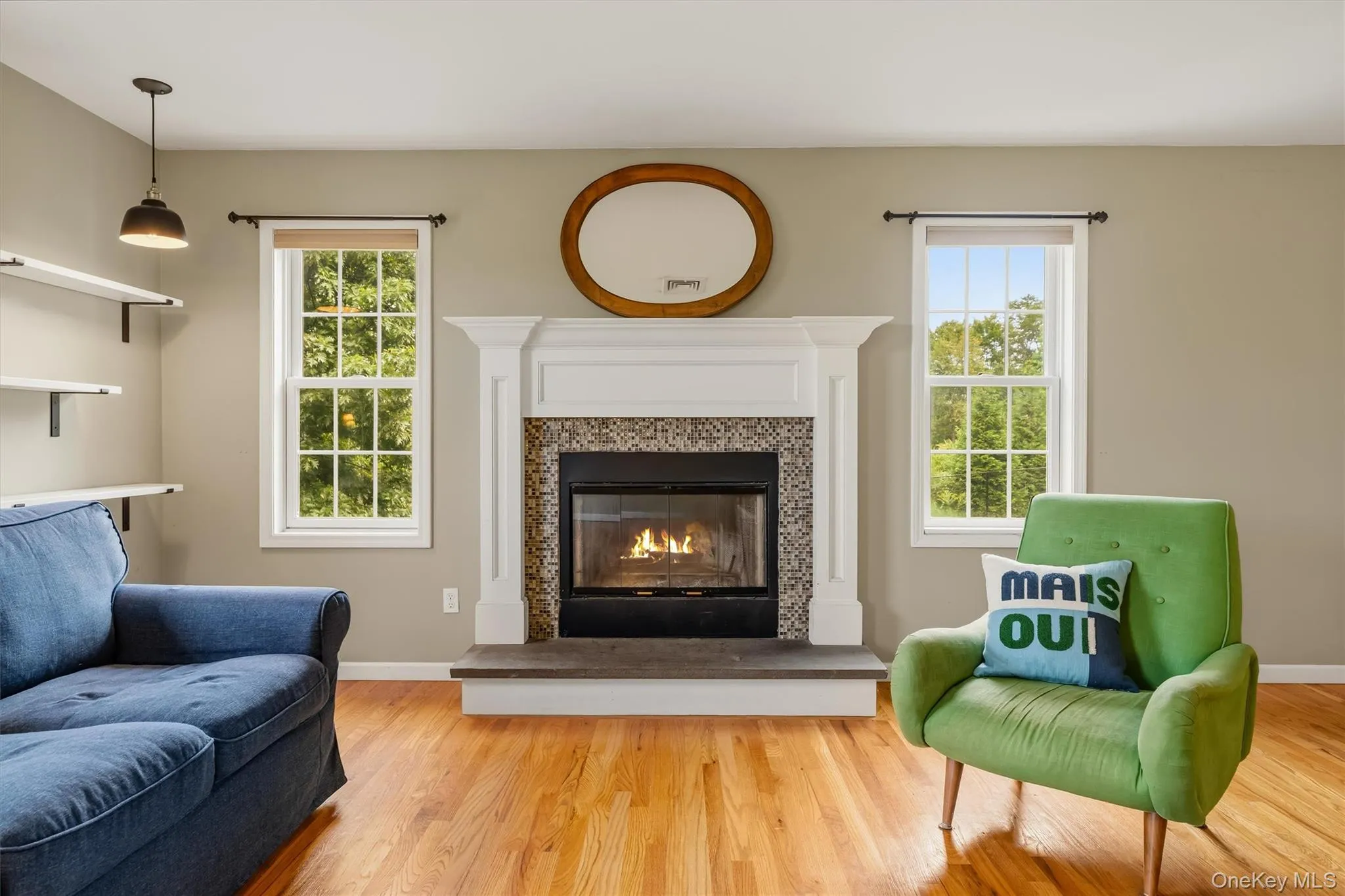 Hardwood flooring, a fireplace with a decorative tile surround and white mantel, two double-hung windows, and a pendant light fixture Hardwood flooring, a fireplace with a decorative tile surround and white mantel, two double-hung windows, and a pendant light fixture