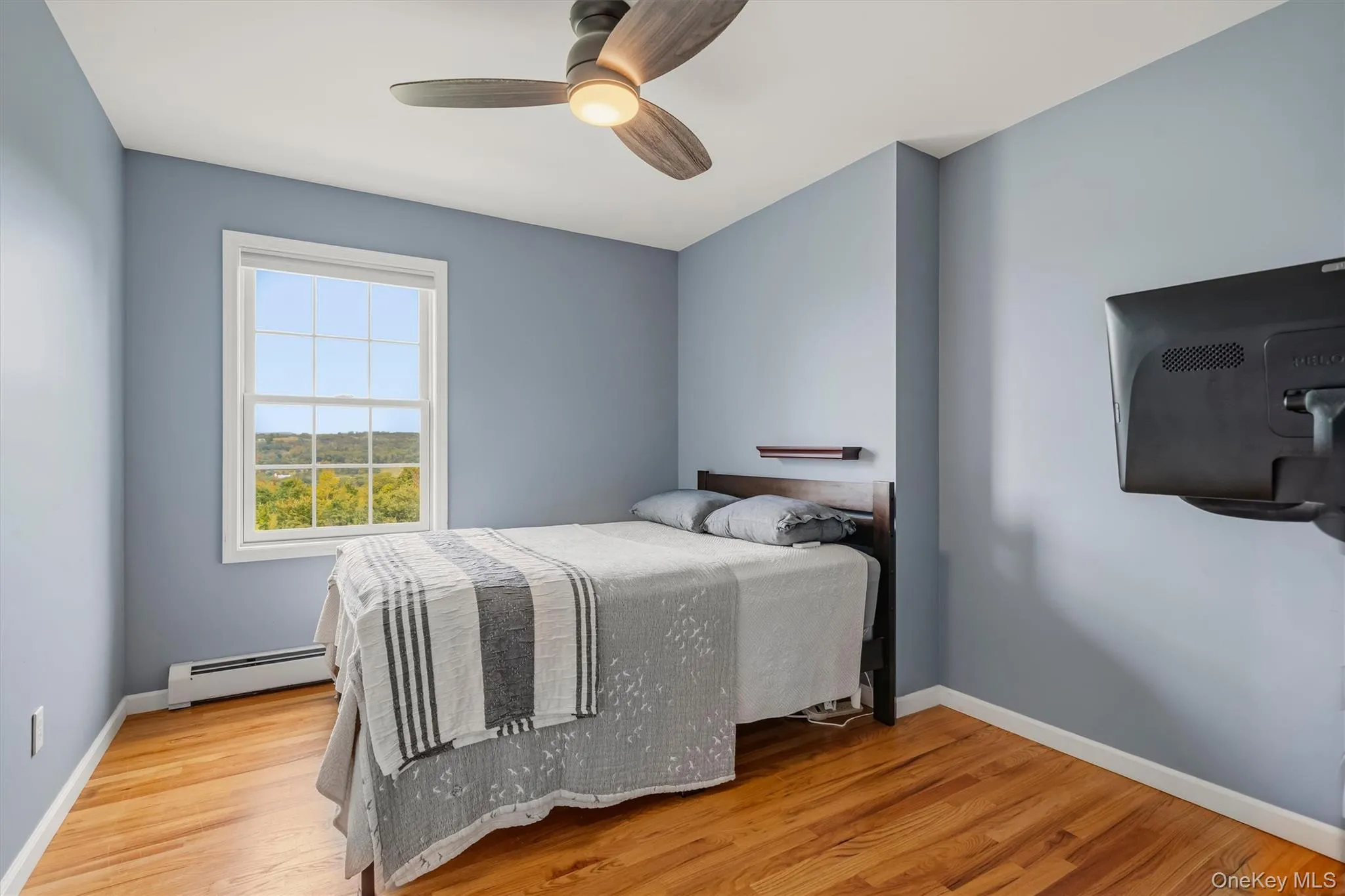 Hardwood flooring, ceiling fan, wall-mounted television bracket, window with exterior scenery, and light blue wall finish Hardwood flooring, ceiling fan, wall-mounted television bracket, window with exterior scenery, and light blue wall finish