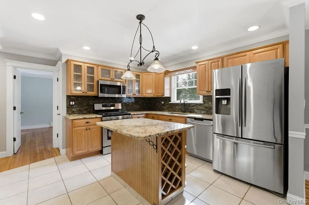 Stainless steel appliances, recessed lighting, central island with built-in wine rack, light wood cabinetry, and a dark stone tile backsplash Stainless steel appliances, recessed lighting, central island with built-in wine rack, light wood cabinetry, and a dark stone tile backsplash