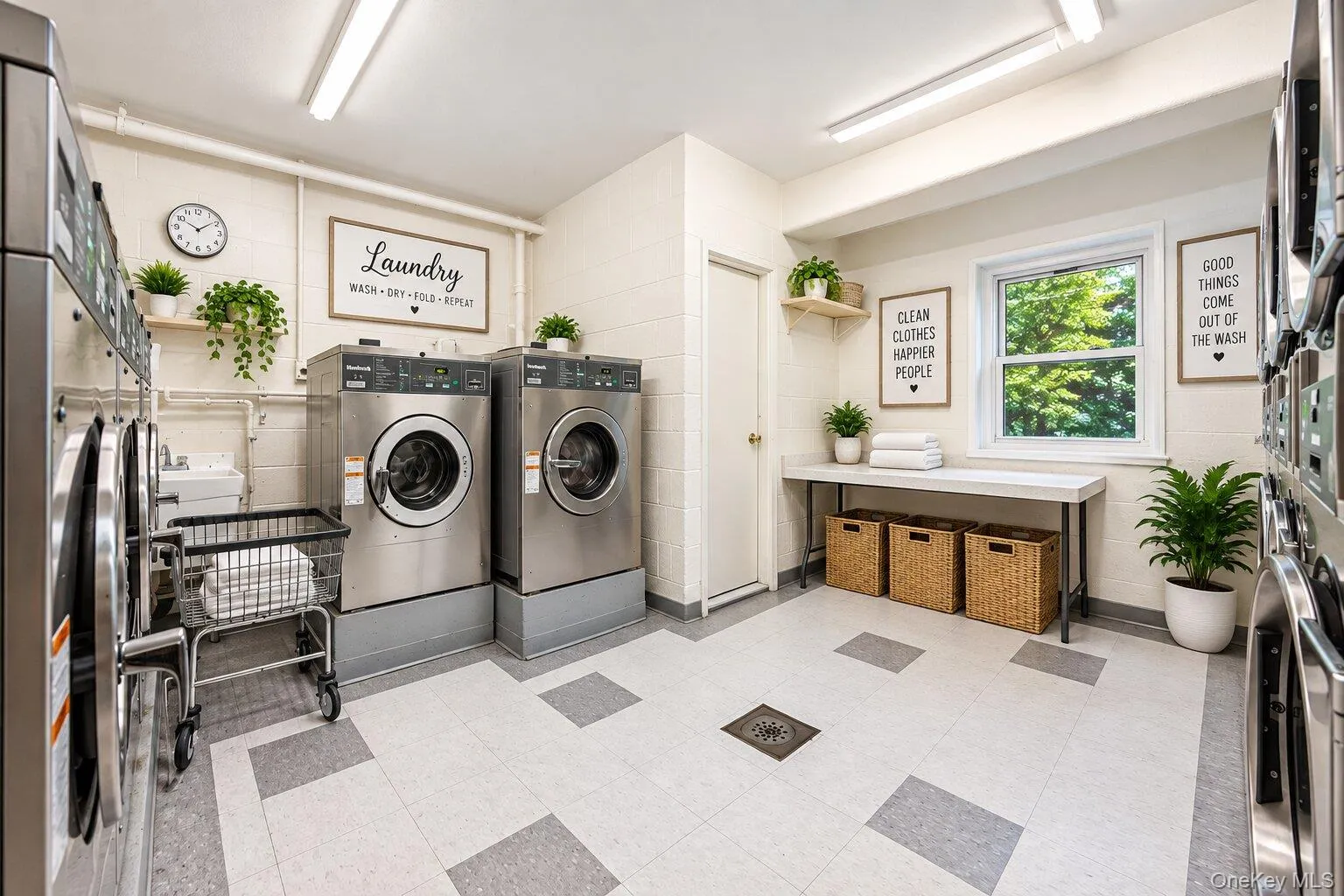 Well-lit laundry facility featuring multiple stainless steel front-load washing machines, a dedicated folding counter with integrated storage, and a large window providing natural light Well-lit laundry facility featuring multiple stainless steel front-load washing machines, a dedicated folding counter with integrated storage, and a large window providing natural light