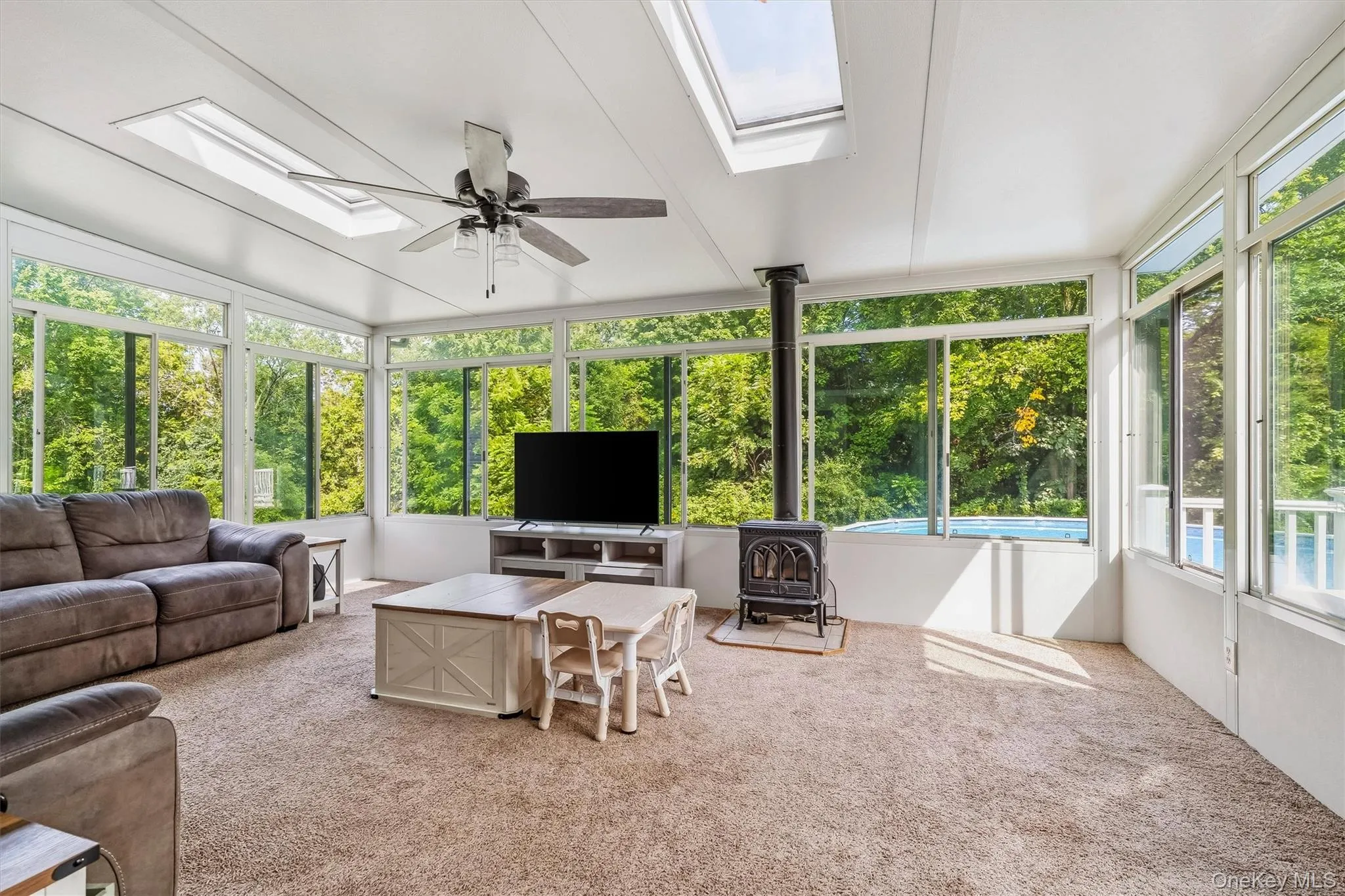 Sunroom featuring abundant natural light, multiple skylights, a ceiling fan, and a wood-burning stove Sunroom featuring abundant natural light, multiple skylights, a ceiling fan, and a wood-burning stove