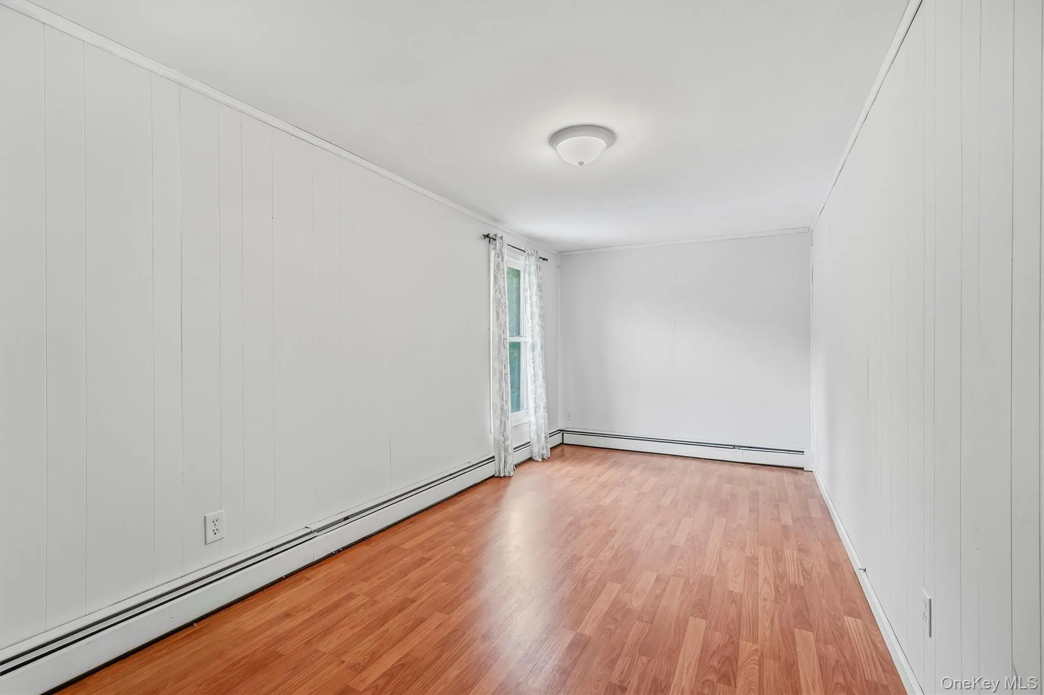 Wood-finish flooring, white paneling on walls, window with trim, and a ceiling-mounted light fixture Wood-finish flooring, white paneling on walls, window with trim, and a ceiling-mounted light fixture