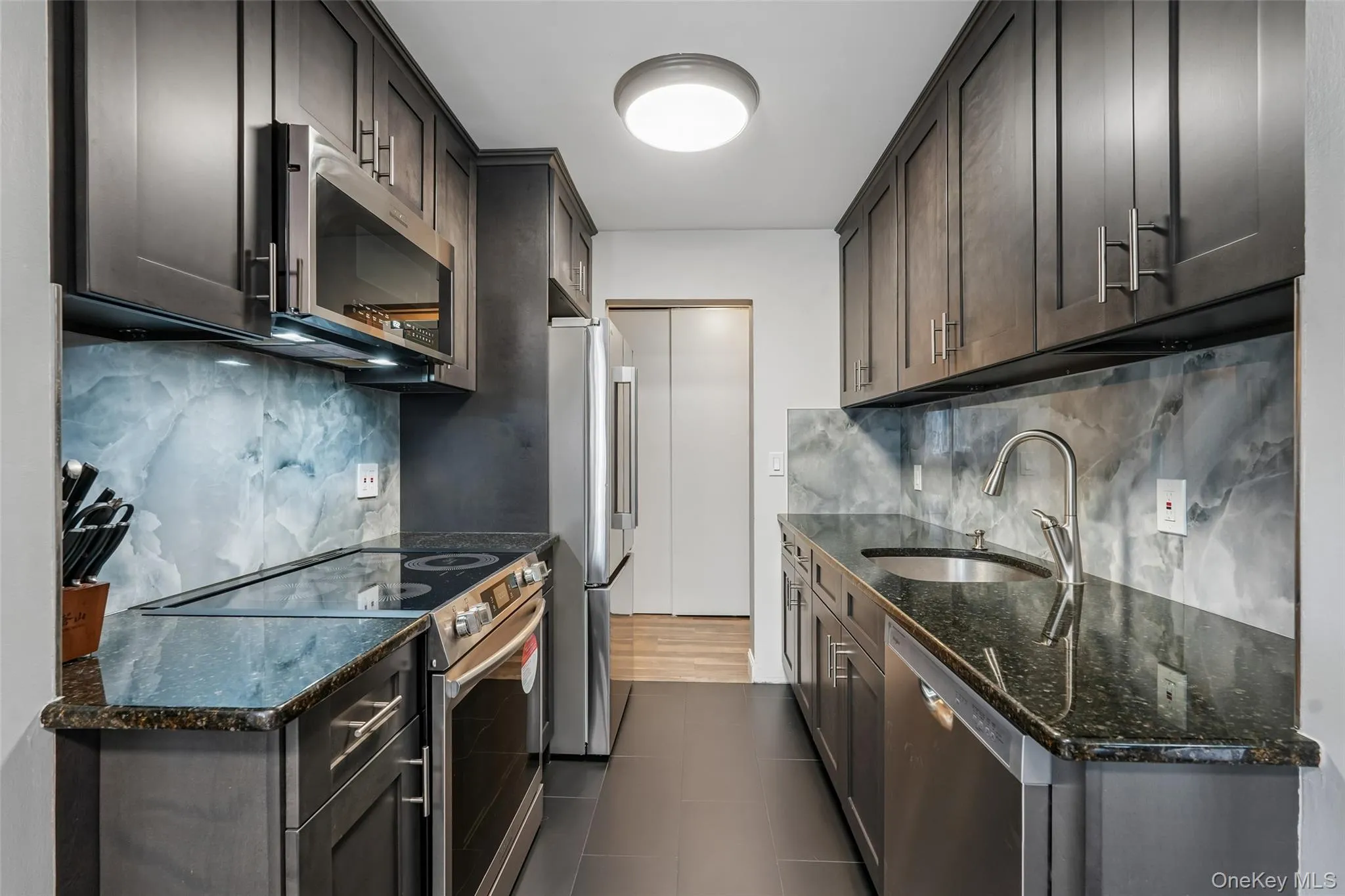 Galley kitchen featuring dark wood-finish cabinetry, stainless steel appliances, granite countertops, and a full-height decorative backsplash Galley kitchen featuring dark wood-finish cabinetry, stainless steel appliances, granite countertops, and a full-height decorative backsplash