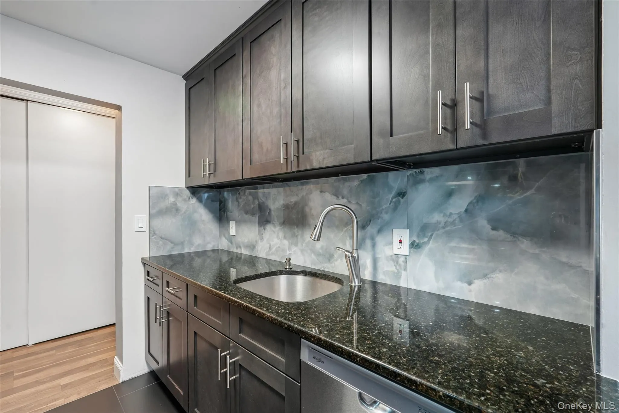 Kitchenette featuring dark wood-finish cabinetry, a stainless steel undermount sink with a gooseneck faucet, a dark speckled countertop, and a patterned backsplash Kitchenette featuring dark wood-finish cabinetry, a stainless steel undermount sink with a gooseneck faucet, a dark speckled countertop, and a patterned backsplash