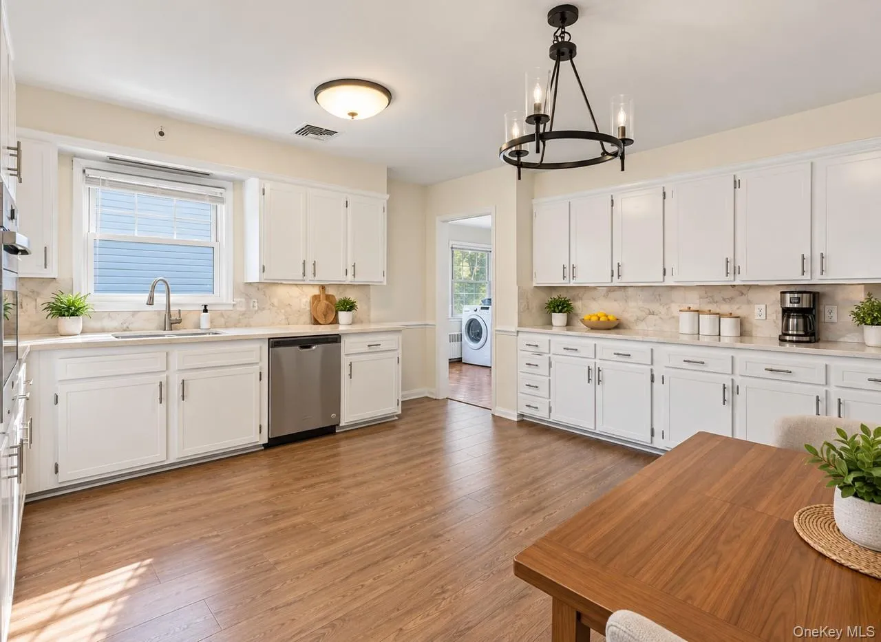 White cabinetry with brushed nickel hardware, light-toned countertops, stainless steel appliances, wood-finish flooring, and a contemporary chandelier White cabinetry with brushed nickel hardware, light-toned countertops, stainless steel appliances, wood-finish flooring, and a contemporary chandelier