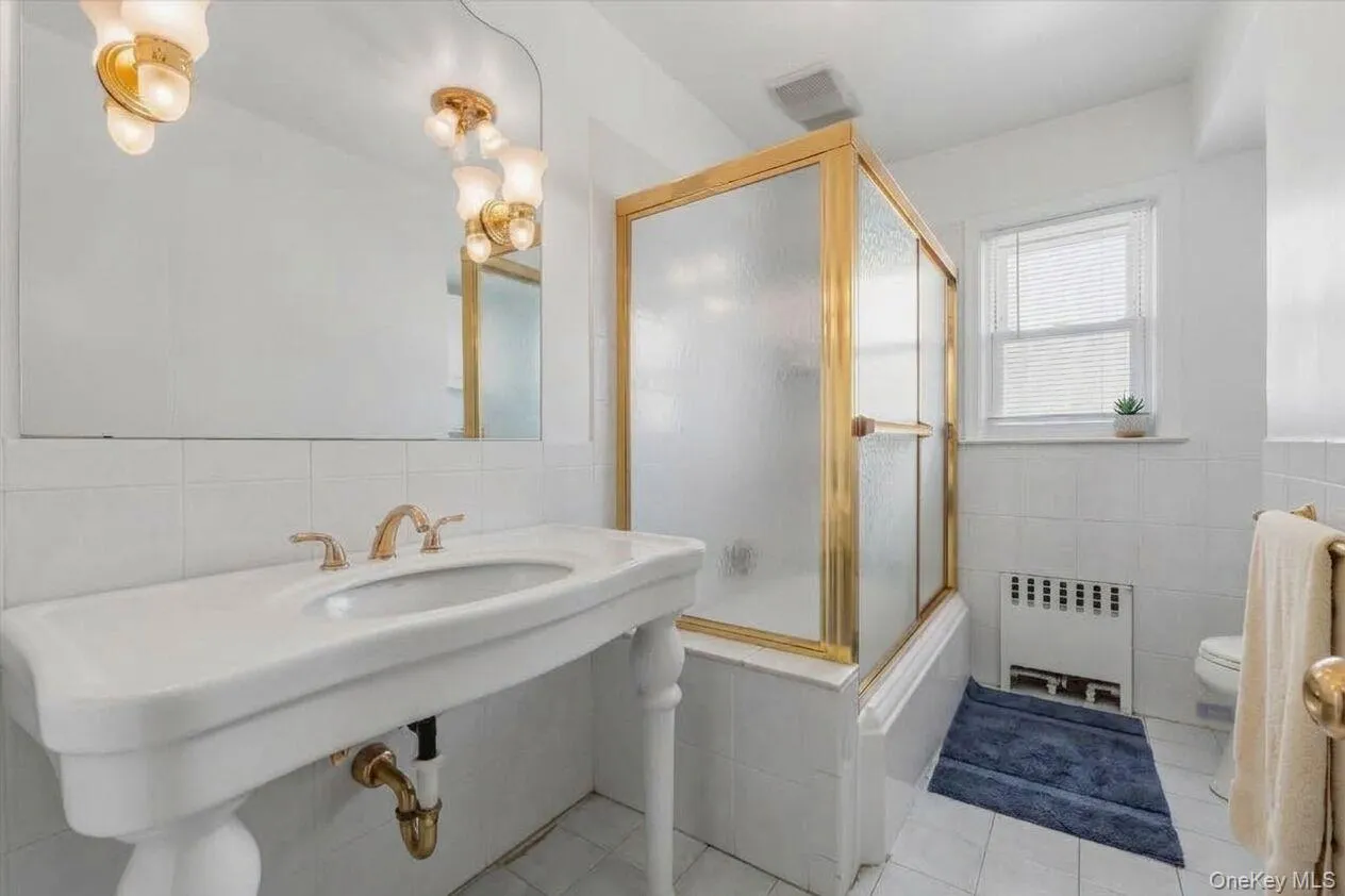Pedestal sink with brass-finish faucet, gold-tone shower enclosure, white tile walls, and suspended lighting fixtures Pedestal sink with brass-finish faucet, gold-tone shower enclosure, white tile walls, and suspended lighting fixtures