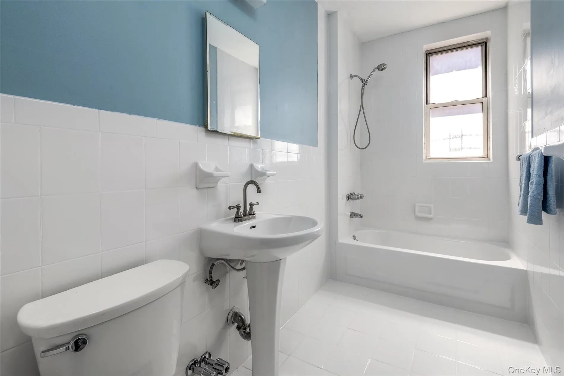 Bathroom featuring a pedestal sink, white ceramic tile walls, a toilet, a bathtub with an integrated shower, and a window Bathroom featuring a pedestal sink, white ceramic tile walls, a toilet, a bathtub with an integrated shower, and a window