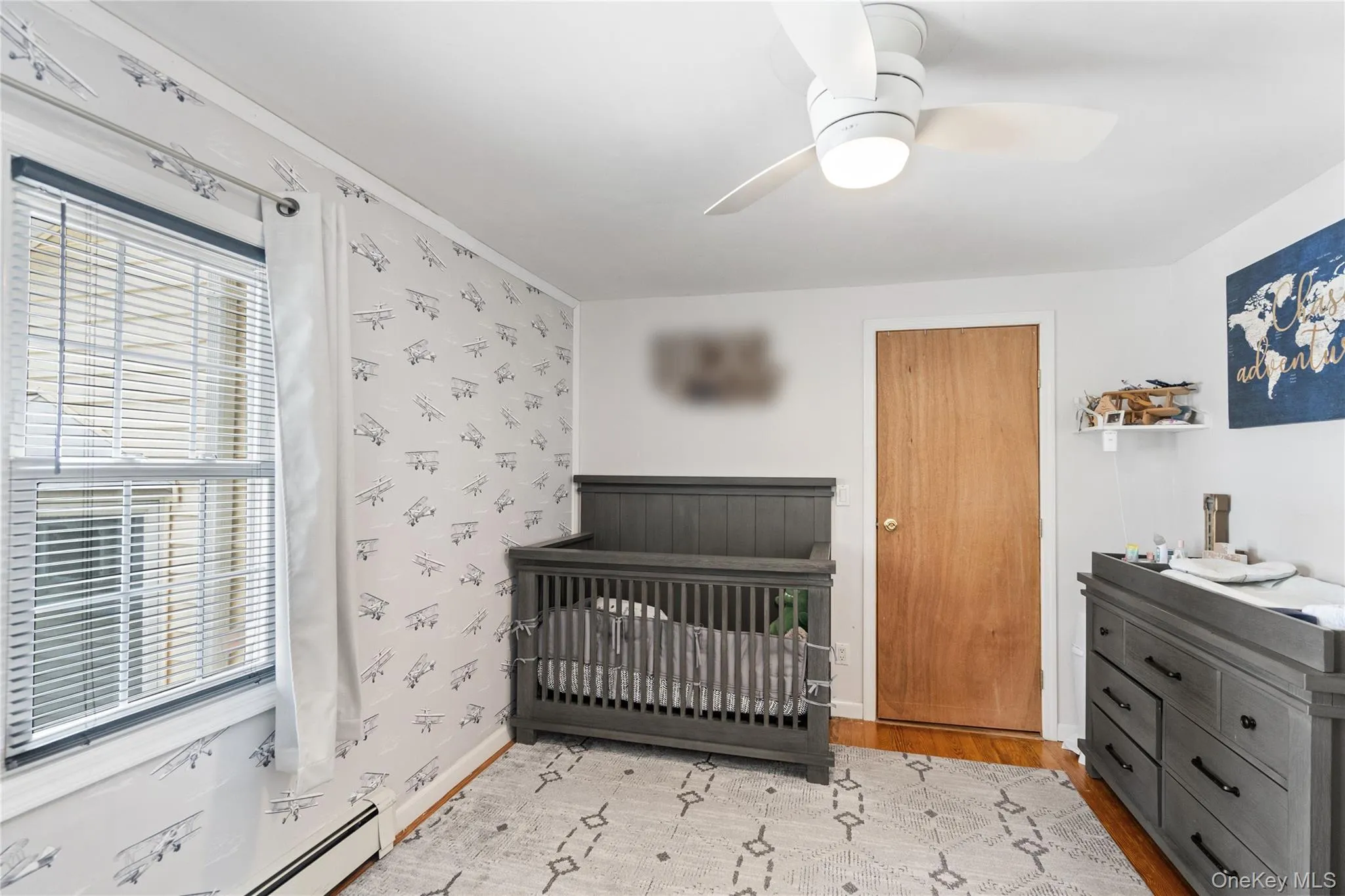 Bedroom featuring light wood finished floors, a crib, ceiling fan, wallpapered walls, and baseboards Bedroom featuring light wood finished floors, a crib, ceiling fan, wallpapered walls, and baseboards