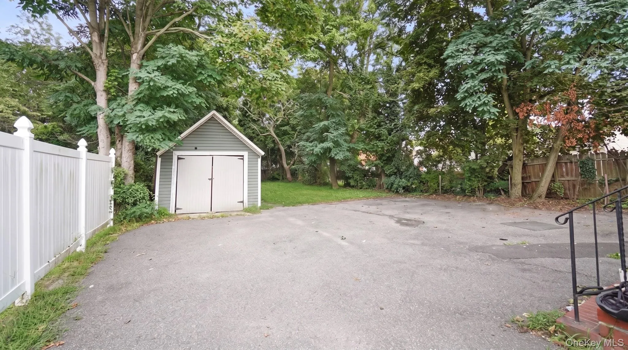 Detached storage shed with panel doors, asphalt driveway, mature trees, and a white privacy fence Detached storage shed with panel doors, asphalt driveway, mature trees, and a white privacy fence