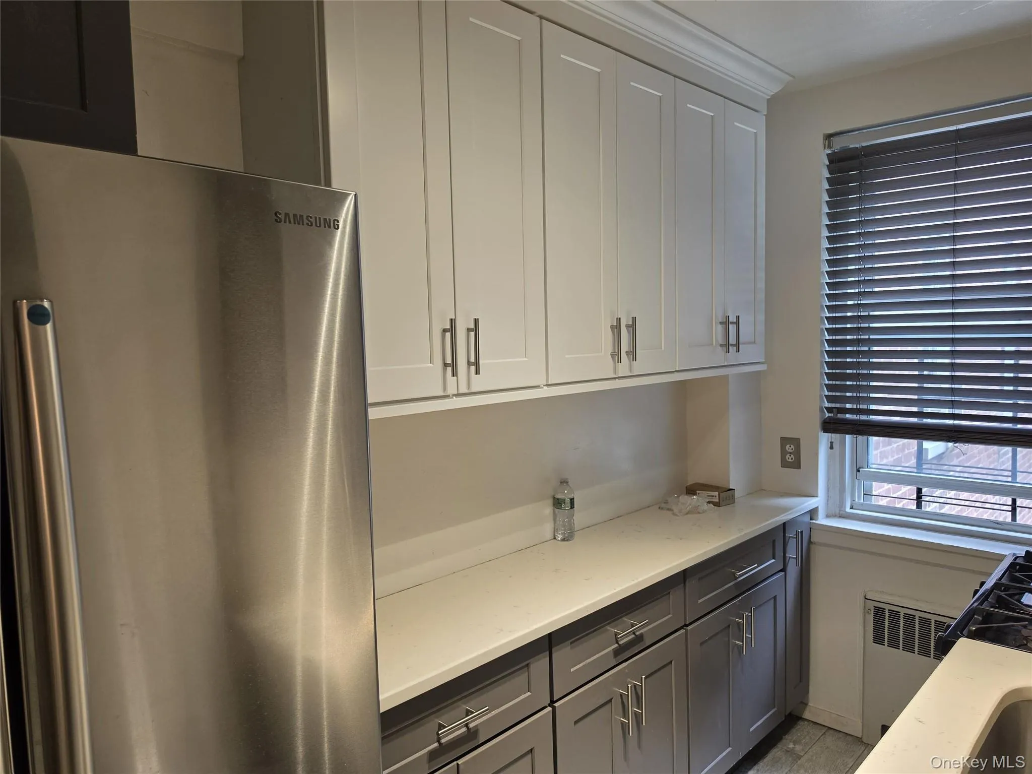 Kitchen featuring stainless steel refrigerator, two-tone cabinetry with upper white shaker-style doors and lower gray shaker-style doors, and light-toned countertops Kitchen featuring stainless steel refrigerator, two-tone cabinetry with upper white shaker-style doors and lower gray shaker-style doors, and light-toned countertops