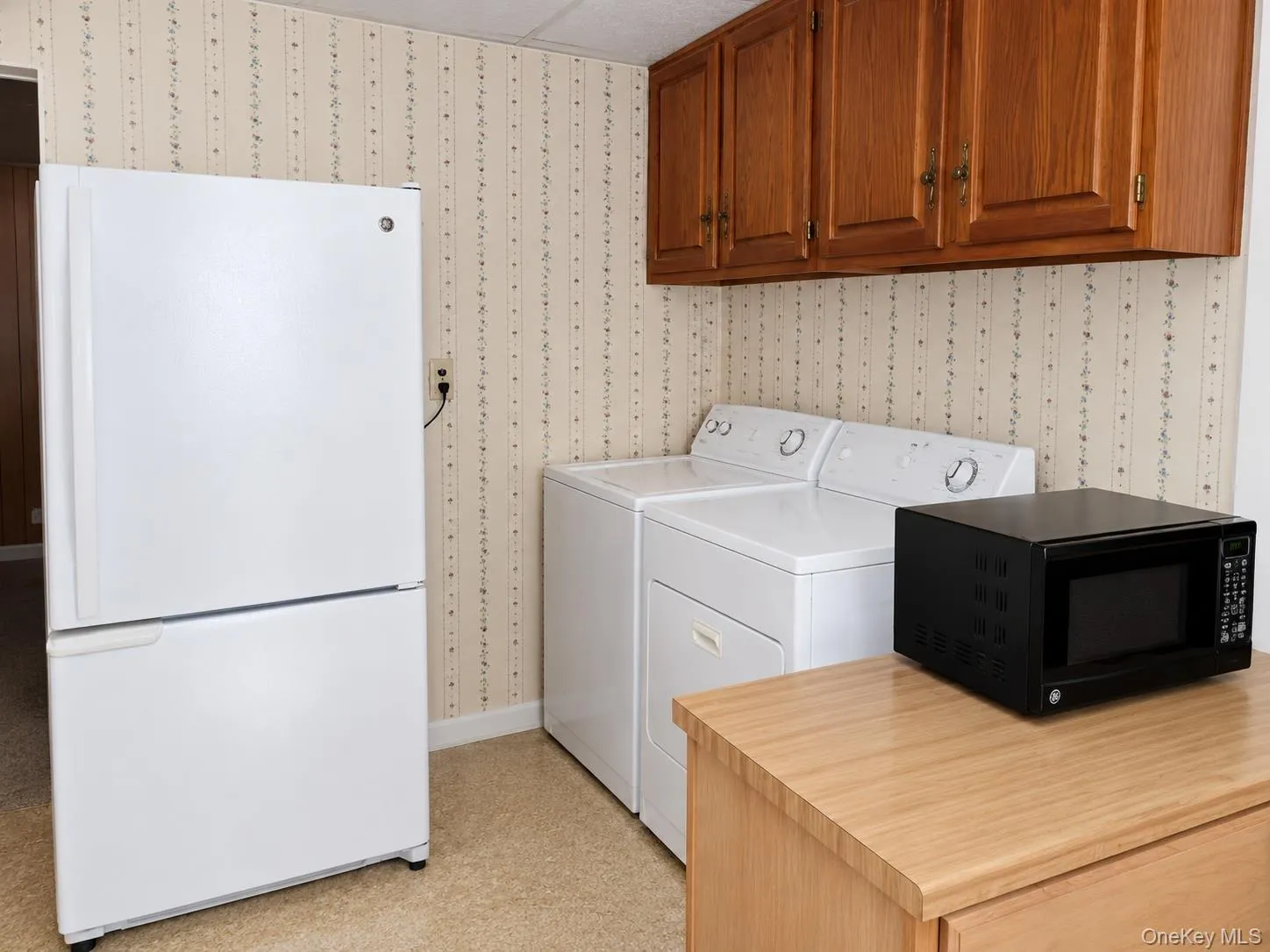 Dedicated utility area featuring wood-finish cabinetry, patterned wallpaper, and light-toned flooring Dedicated utility area featuring wood-finish cabinetry, patterned wallpaper, and light-toned flooring