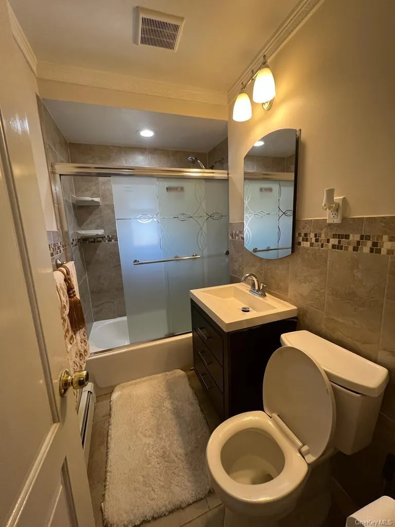 Tiled bathroom featuring a glass-enclosed shower with a tub, dark wood-finish vanity with an integrated sink, and an arched wall mirror Tiled bathroom featuring a glass-enclosed shower with a tub, dark wood-finish vanity with an integrated sink, and an arched wall mirror