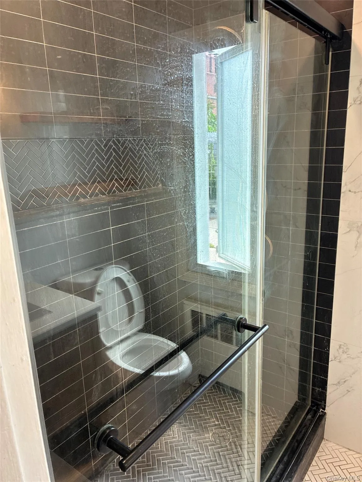 Glass-enclosed shower with black-framed door, dark subway tile walls, and a herringbone tile accent band Glass-enclosed shower with black-framed door, dark subway tile walls, and a herringbone tile accent band