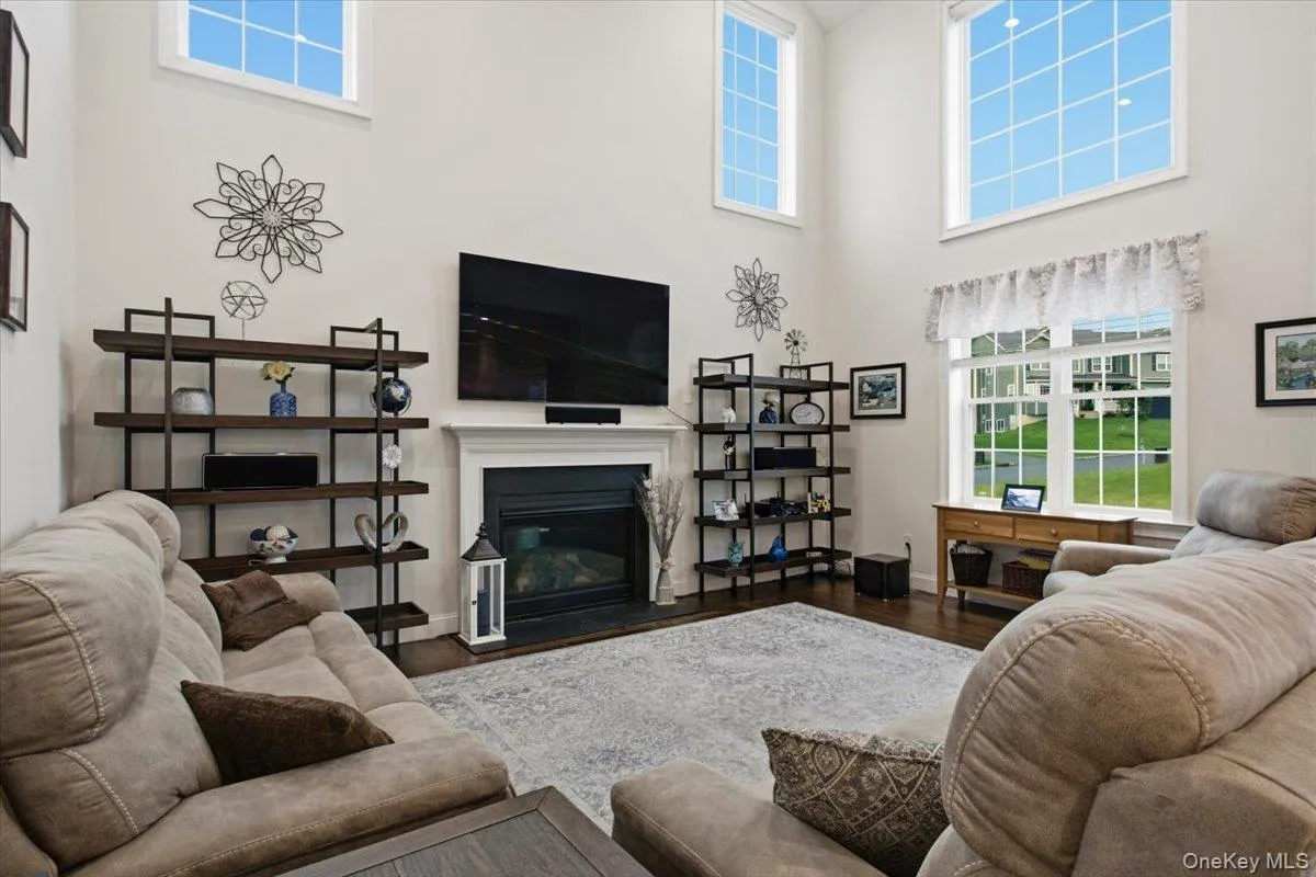 Two-story living area featuring a fireplace with a white mantel, wood-finish flooring, built-in shelving, and multiple large windows Two-story living area featuring a fireplace with a white mantel, wood-finish flooring, built-in shelving, and multiple large windows