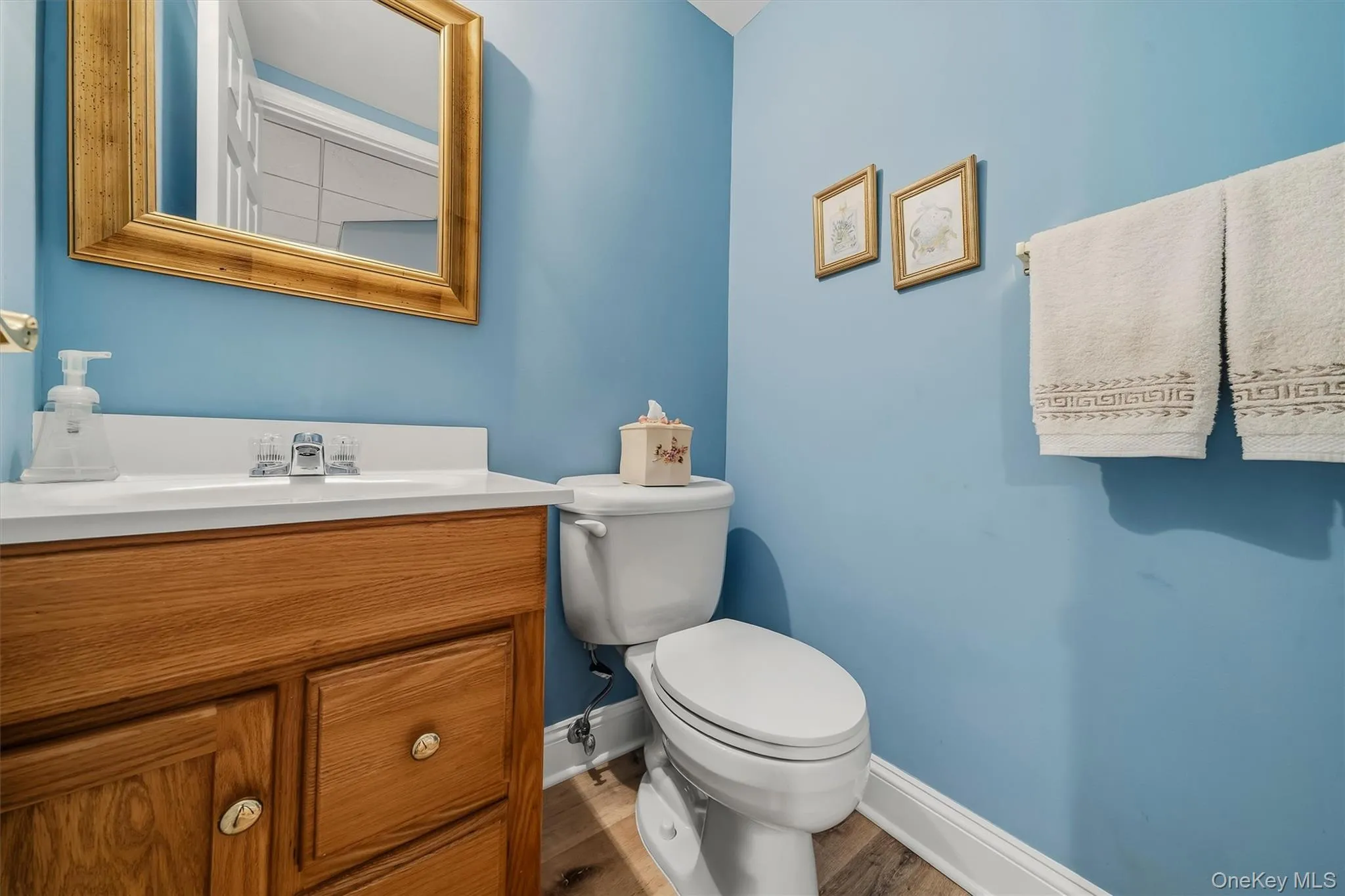 Wood-finish flooring, light wood vanity with brass hardware, white countertop with integrated sink, chrome faucet, and a toilet Wood-finish flooring, light wood vanity with brass hardware, white countertop with integrated sink, chrome faucet, and a toilet