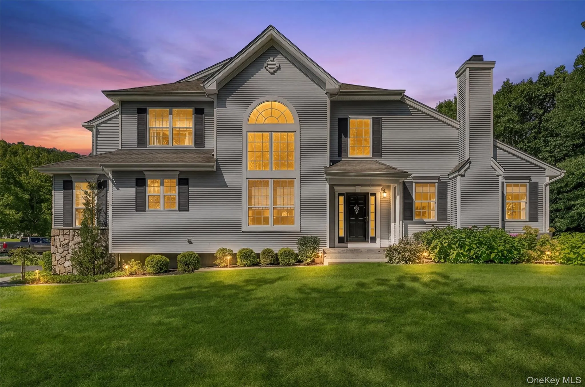VIRTUAL STAGING Gray siding exterior with black shutters, prominent arched window, stone accent column, front entry with transom window, and established perimeter landscaping VIRTUAL STAGING Gray siding exterior with black shutters, prominent arched window, stone accent column, front entry with transom window, and established perimeter landscaping
