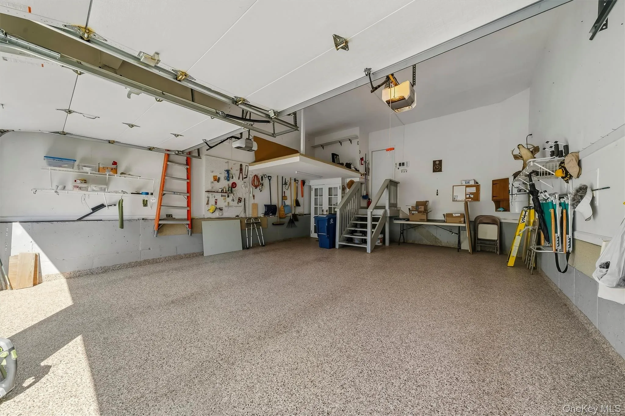 Spacious garage with speckled epoxy flooring, overhead door openers, built-in shelving, and a raised storage platform with staircase access Spacious garage with speckled epoxy flooring, overhead door openers, built-in shelving, and a raised storage platform with staircase access