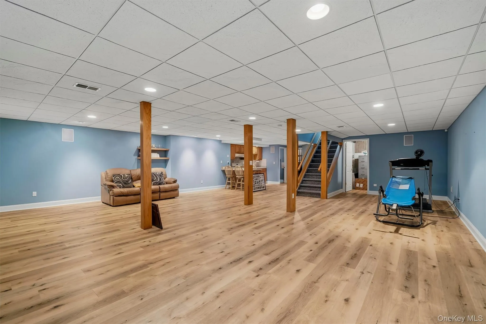 Wood-finish flooring, recessed lighting, drop ceiling, light blue walls, and a visible staircase Wood-finish flooring, recessed lighting, drop ceiling, light blue walls, and a visible staircase