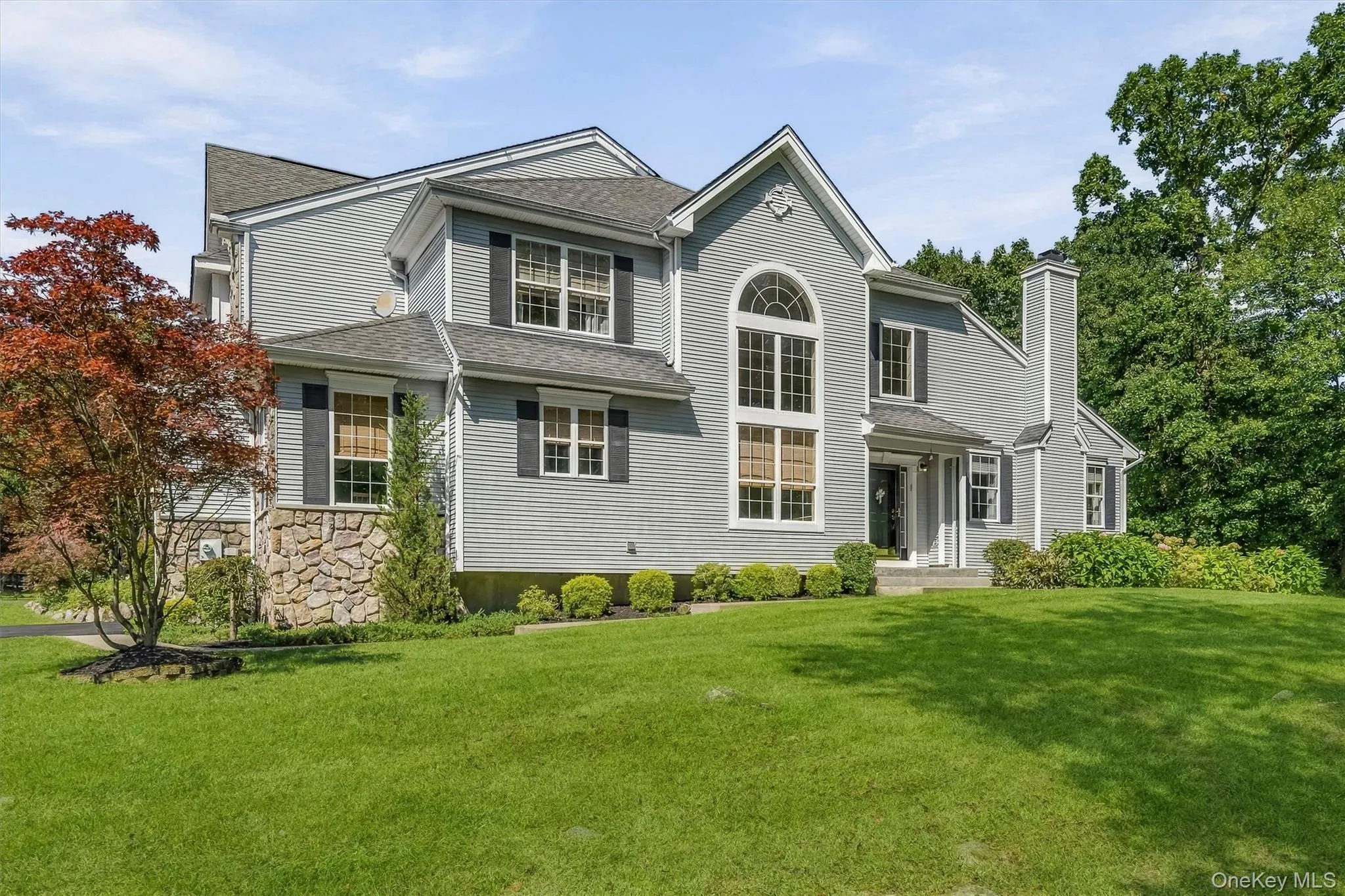 Expansive front lawn with mature landscaping, a stone veneer foundation, multiple windows with black shutters, an arched transom window, and a prominent chimney Expansive front lawn with mature landscaping, a stone veneer foundation, multiple windows with black shutters, an arched transom window, and a prominent chimney