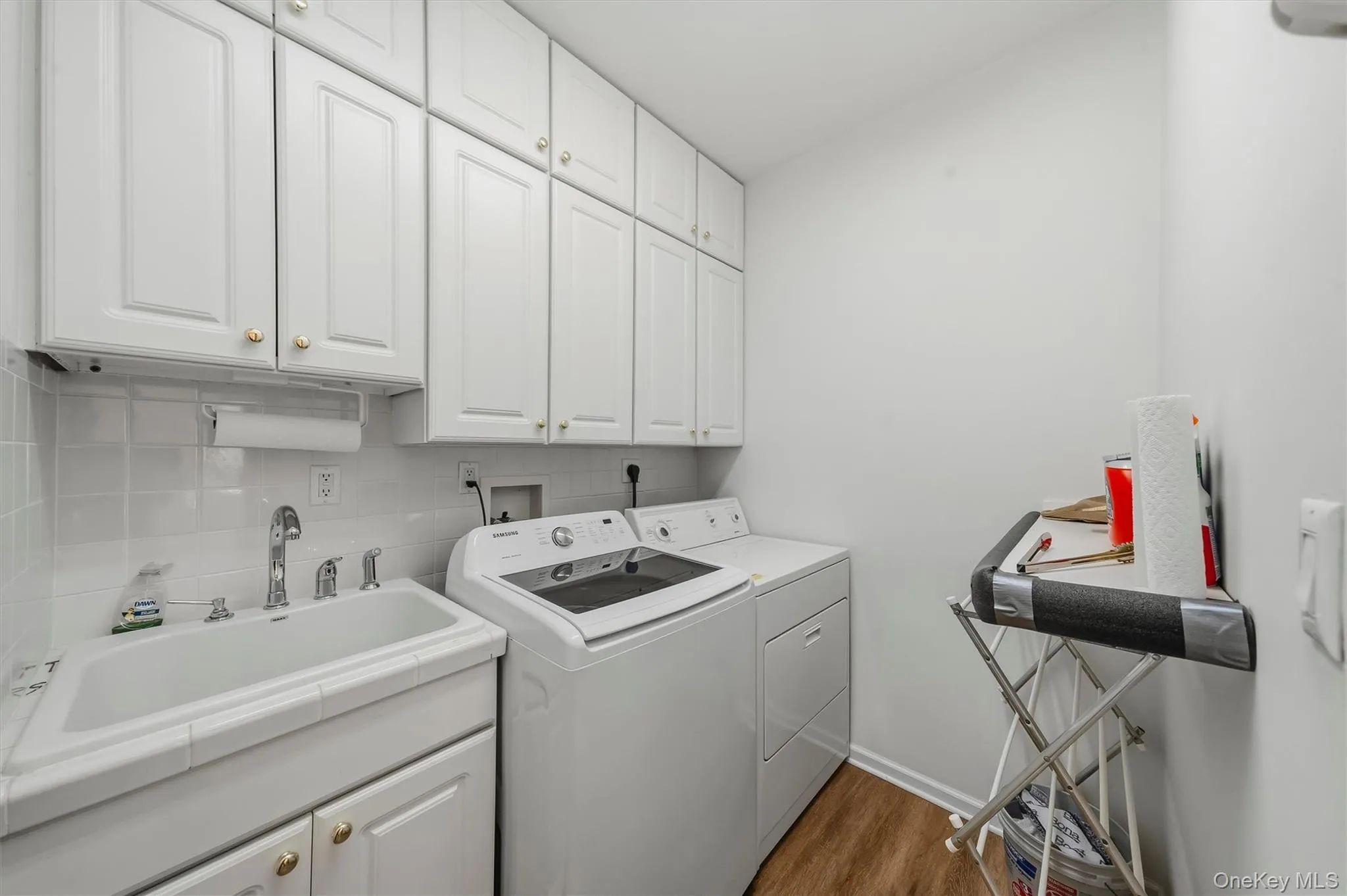 White cabinetry with gold-tone hardware, utility sink with chrome faucet, white square tile backsplash, wood-finish flooring White cabinetry with gold-tone hardware, utility sink with chrome faucet, white square tile backsplash, wood-finish flooring