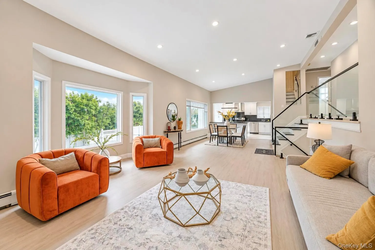 Open-concept living area featuring a vaulted ceiling with recessed lighting, bay windows, light wood-finish flooring, and a modern staircase with glass railing Open-concept living area featuring a vaulted ceiling with recessed lighting, bay windows, light wood-finish flooring, and a modern staircase with glass railing
