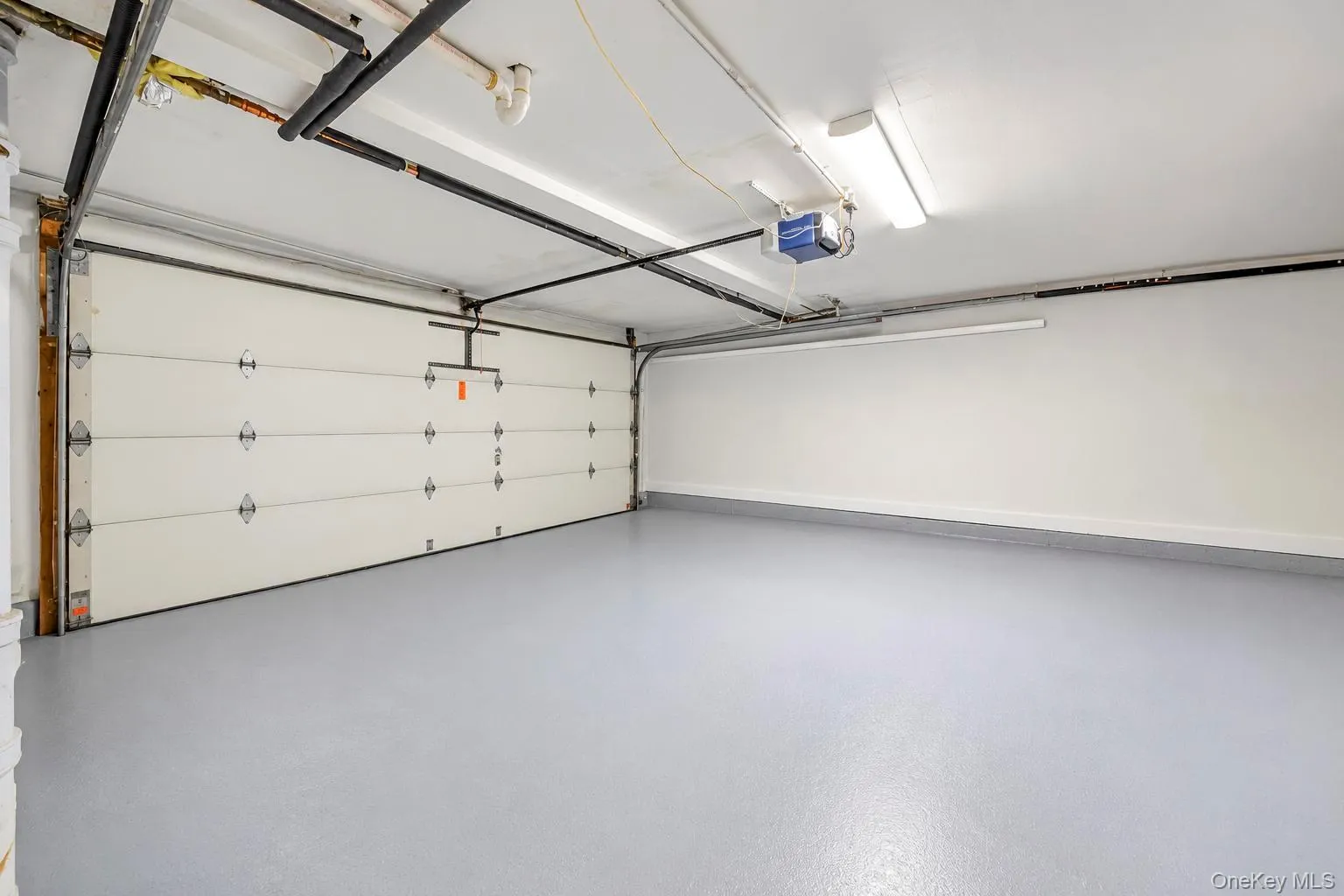 Spacious garage with a light gray epoxy floor coating, white walls, overhead fluorescent lighting, and a white paneled garage door with an automatic opener Spacious garage with a light gray epoxy floor coating, white walls, overhead fluorescent lighting, and a white paneled garage door with an automatic opener