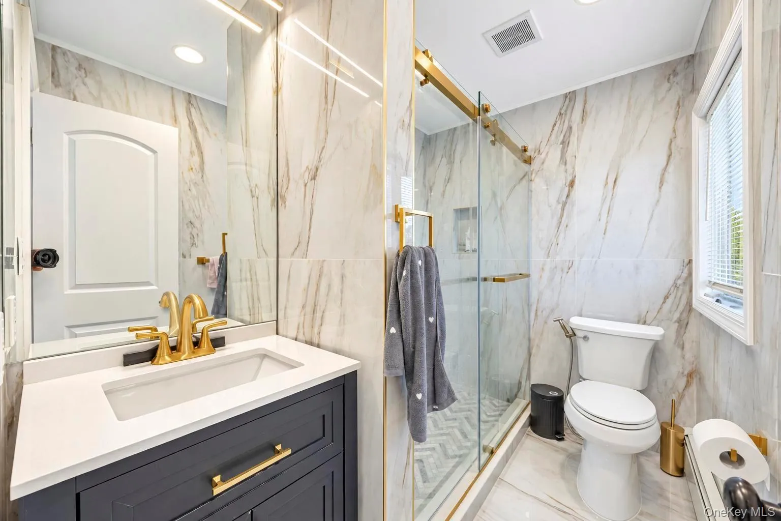 Marble-look wall tile, glass-enclosed shower with gold-tone fixtures, white vanity countertop, gold-tone sink faucet, and built-in vanity cabinetry Marble-look wall tile, glass-enclosed shower with gold-tone fixtures, white vanity countertop, gold-tone sink faucet, and built-in vanity cabinetry